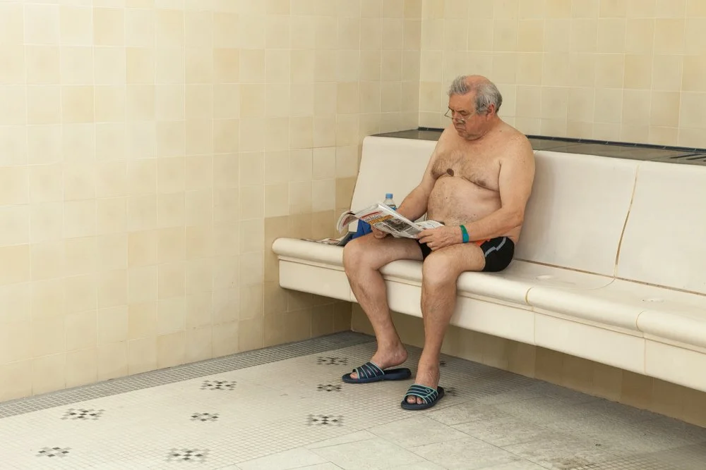 Un homme âgé assis seul sur un banc dans une pièce carrelée, lit un journal, portant des chaussons et un bracelet bleu.