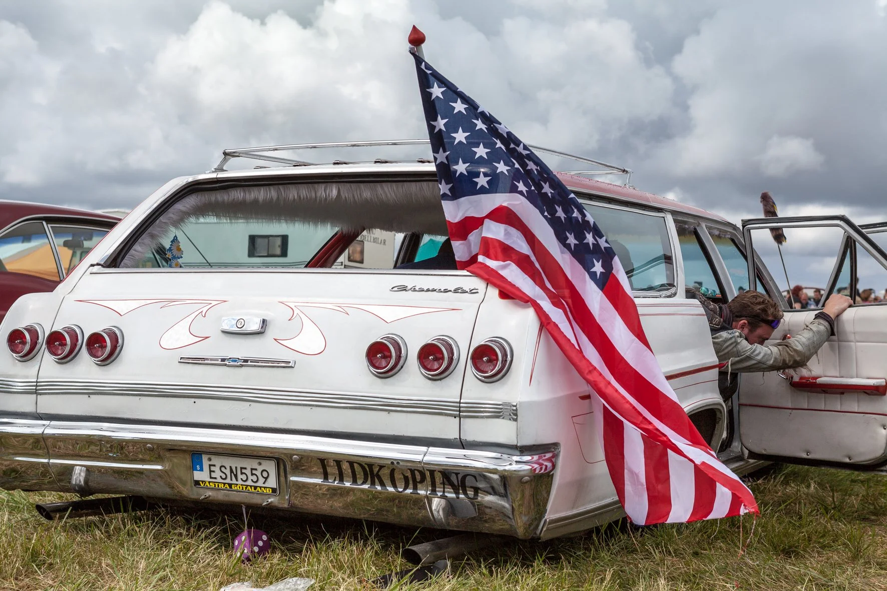 Voiture vintage blanche avec un drapeau américain attaché à l'arrière, stationnée sur de l'herbe, avec un homme qui vomit à l'extérieur du véhicule, sous un ciel nuageux.