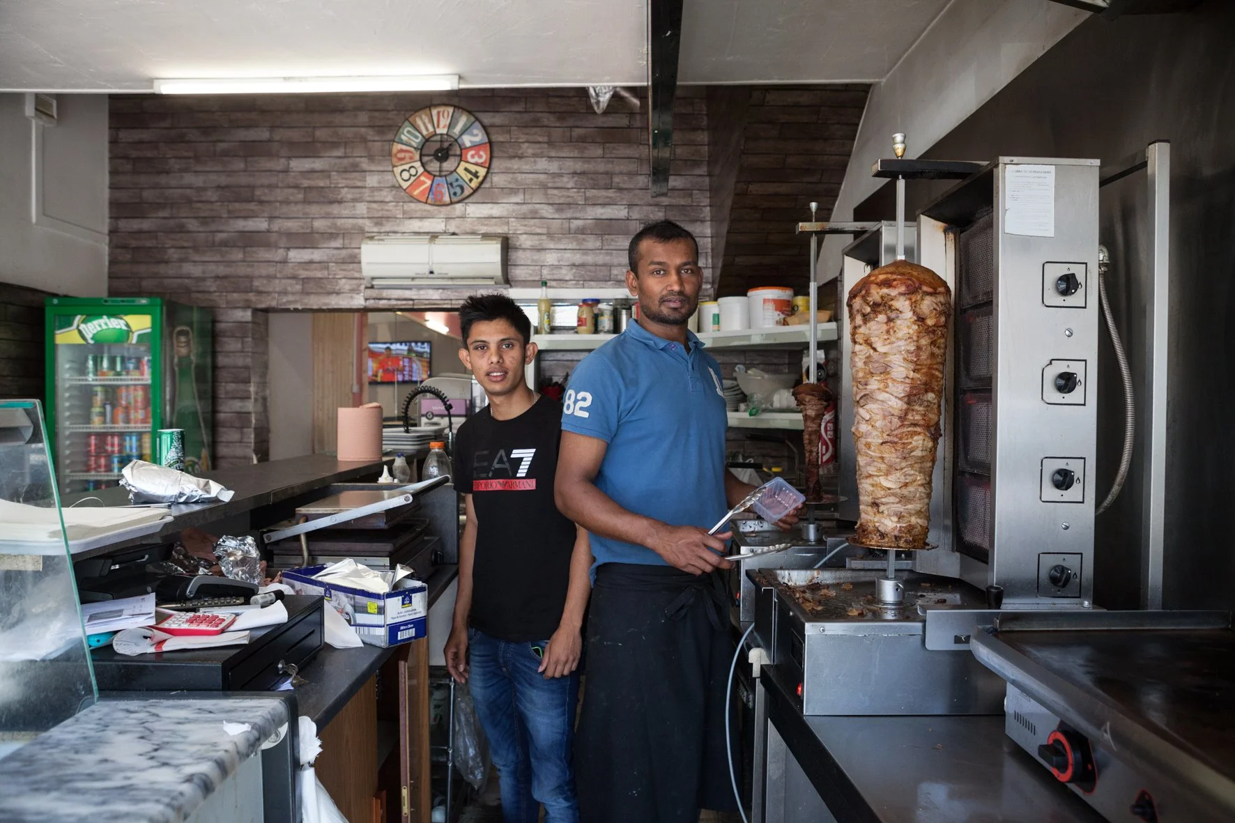 Deux hommes dans une cuisine, l'un en tee-shirt noir, l'autre en polo bleu, préparant un kebab sur une machine à cuire la viande.