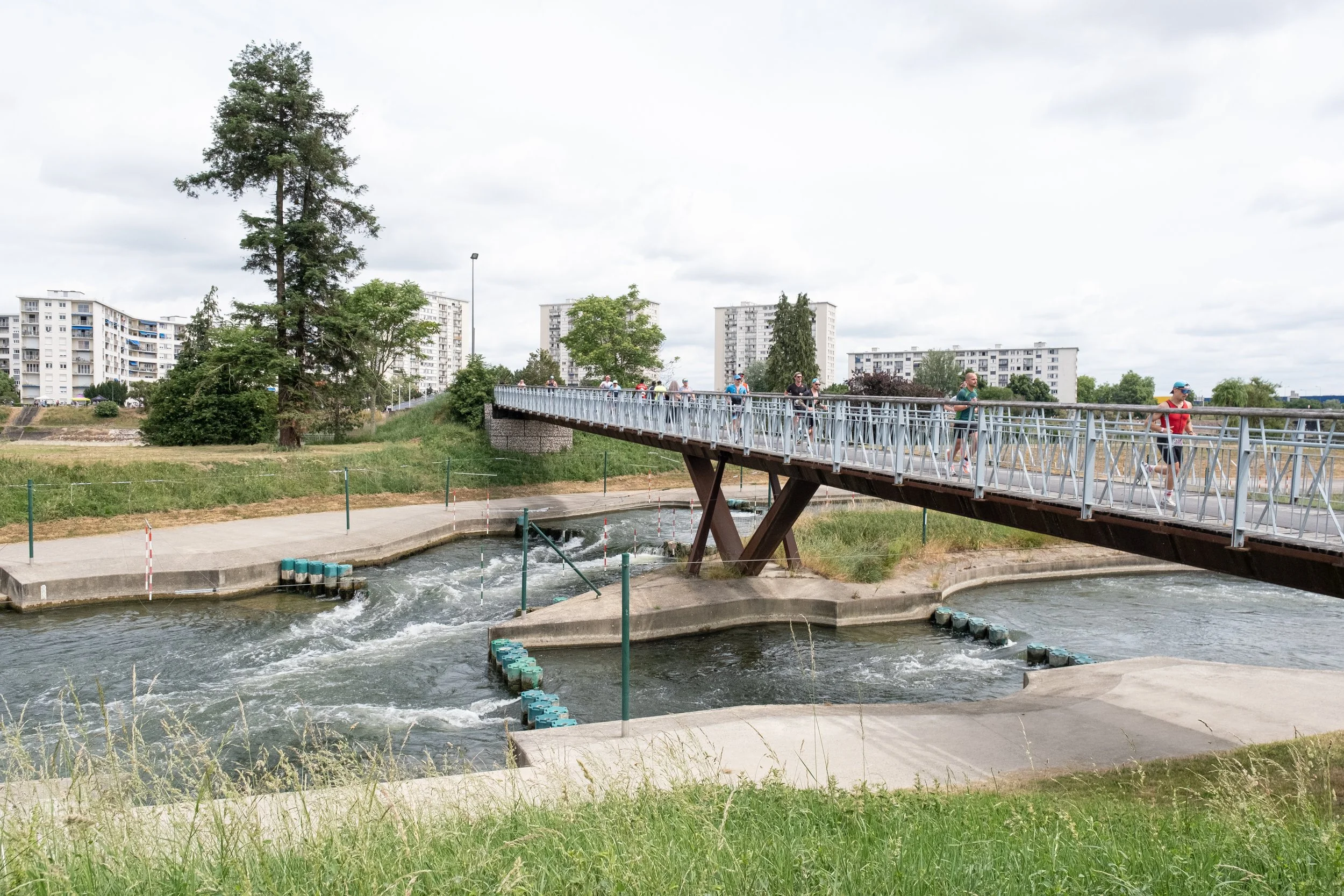Un pont piétonnier en métal traverse une rivière en milieu urbain à Tours, avec plusieurs personnes marchant dessus, des arbres et des immeubles résidentiels en arrière-plan, ciel nuageux.