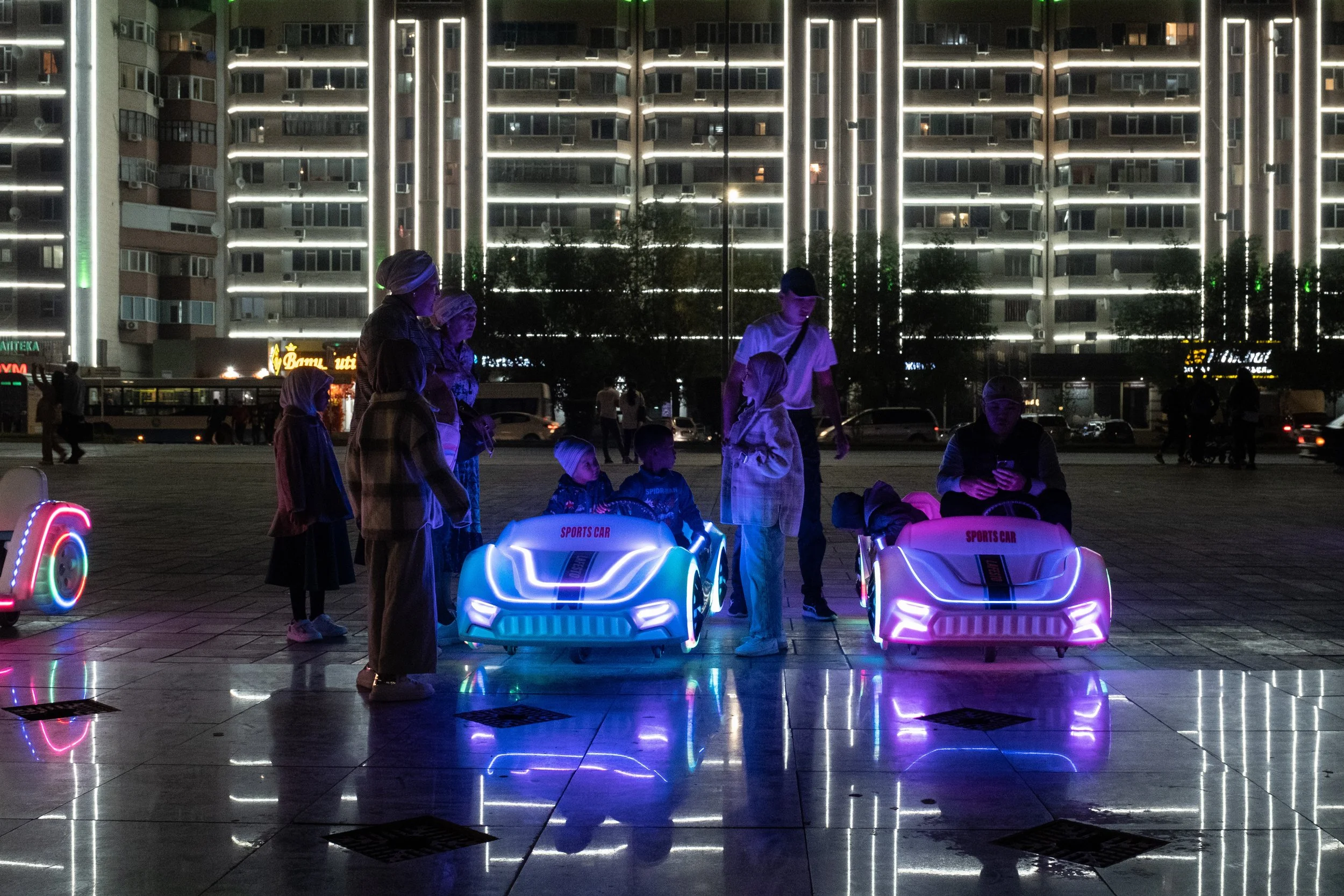 Enfants jouant en conduisant des voitures électriques illuminées dans un espace public la nuit, avec un bâtiment éclairé en arrière-plan, au Kazakhstan.