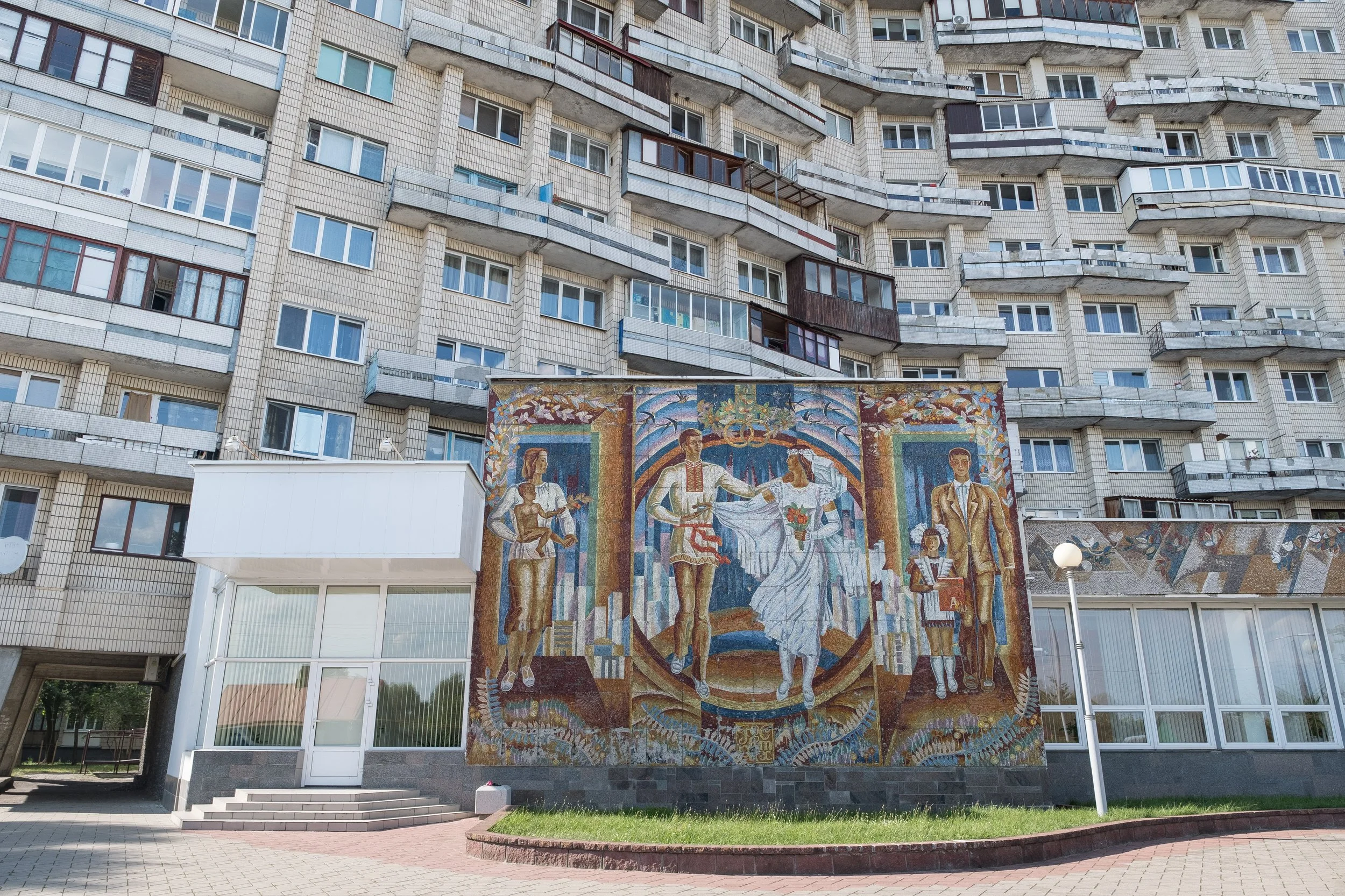 Mosaïque murale soviétique représentant une scène de mariage avec un couple, entourés de deux enfants et d'autres personnes, au pied d'un immeuble résidentiel en Biélorussie.