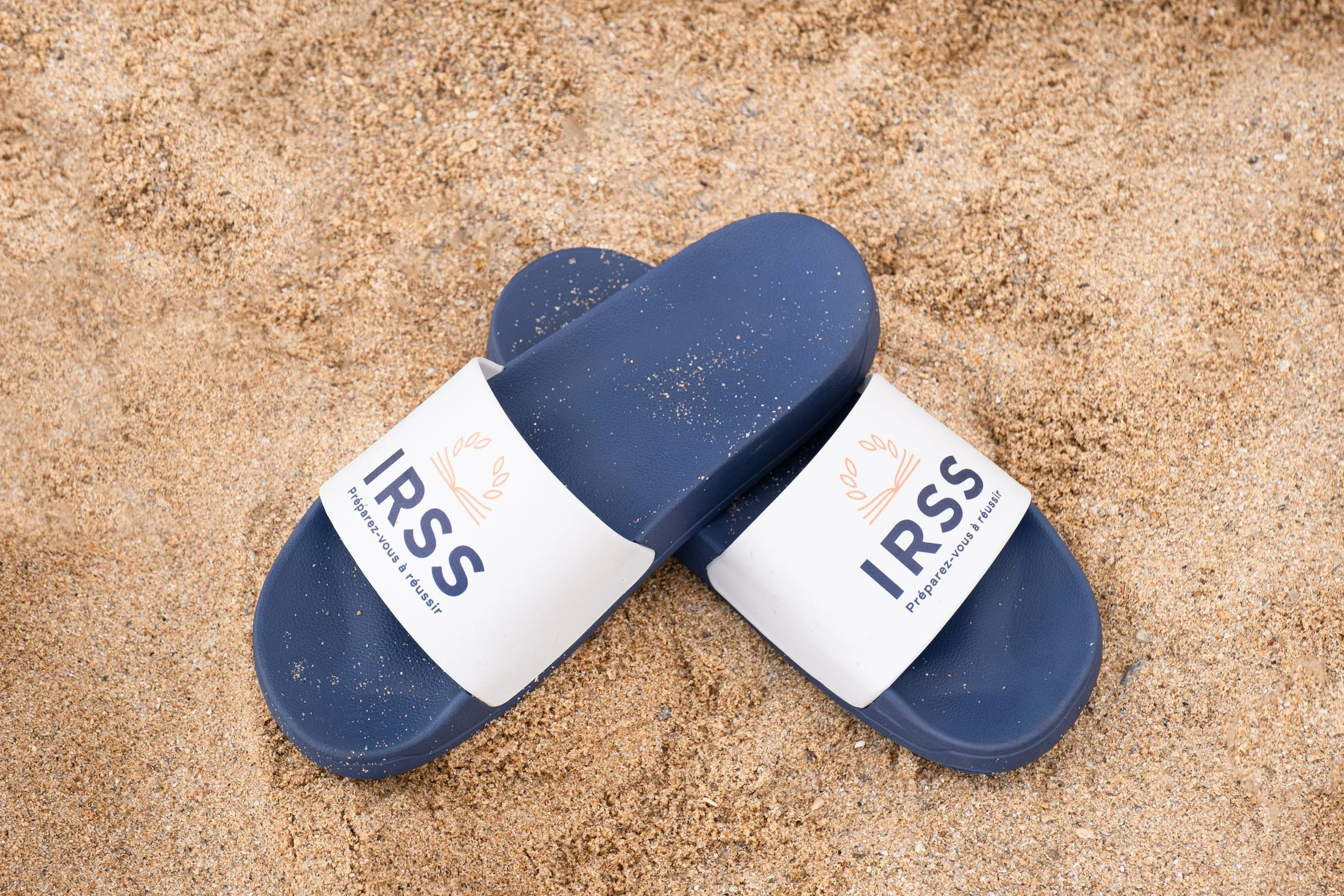 Une paire de tongs bleues avec une bande blanche portant le texte 'IRSS' et un slogan en français, posées sur du sable.