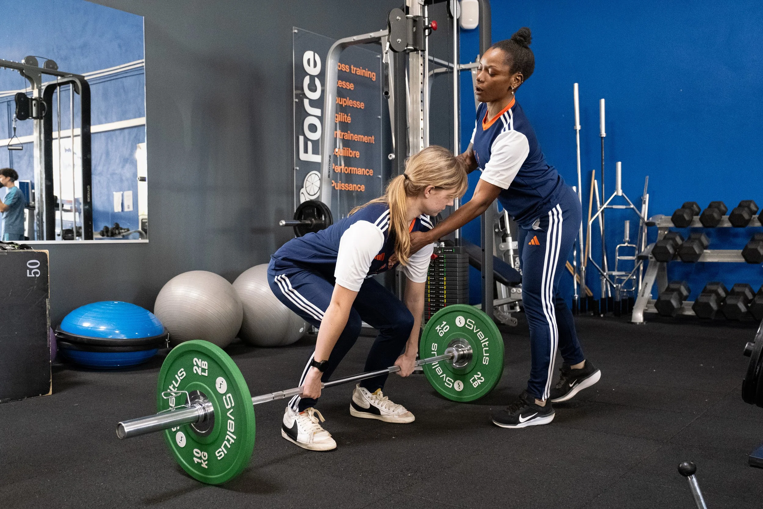 Une femme aide une autre femme à soulever une barre avec des poids dans une salle de sport.