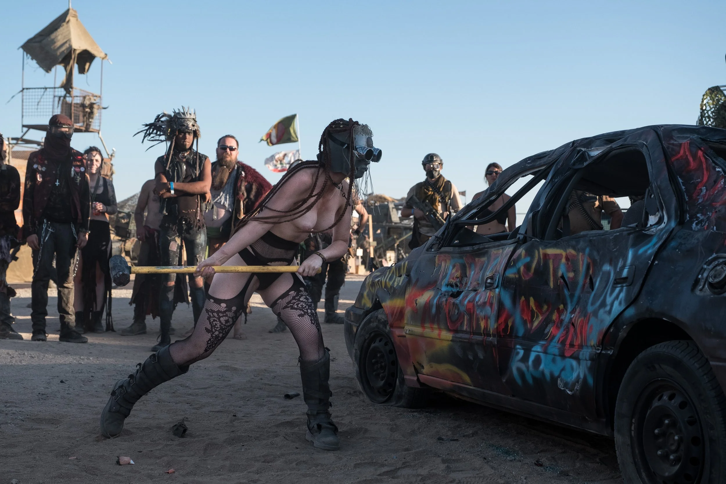 Une femme portant un masque et des lunettes de vision nocturne casse une voiture avec un marteau, entourée de plusieurs personnes costumées dans le désert du Nevada à Wasteland.