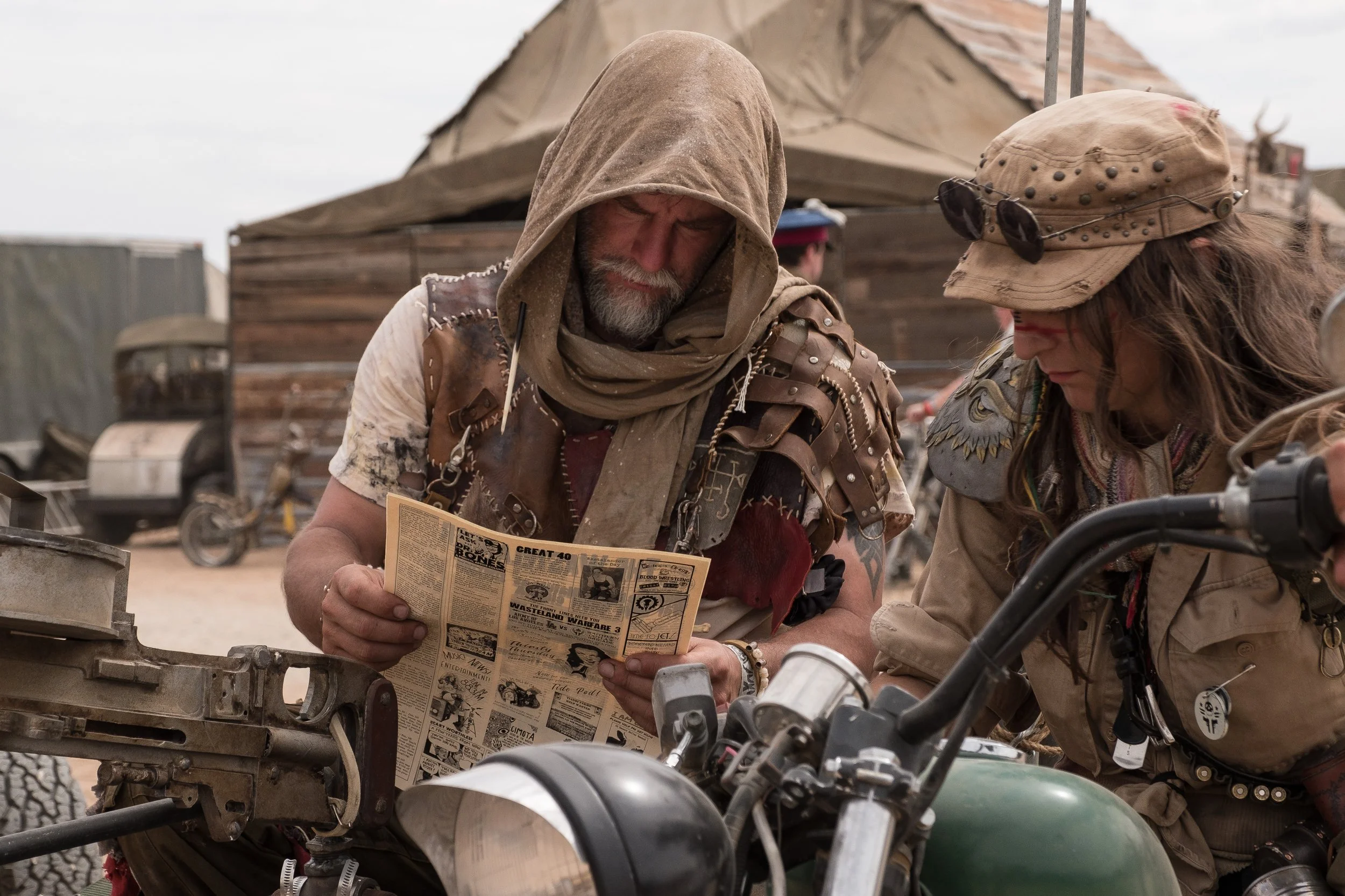 Deux personnes en tenue post-apocalyptique regardant un journal, avec une moto en premier plan et un campement en arrière-plan à Wasteland.