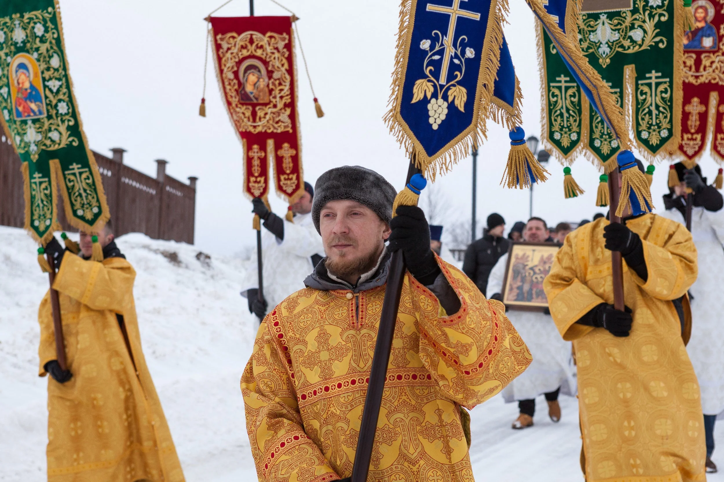 Groupe de personnes participant à une procession religieuse en hiver, portant des icônes et des bannières avec des motifs religieux, vêtus de vêtements traditionnels dorés et ornementés.