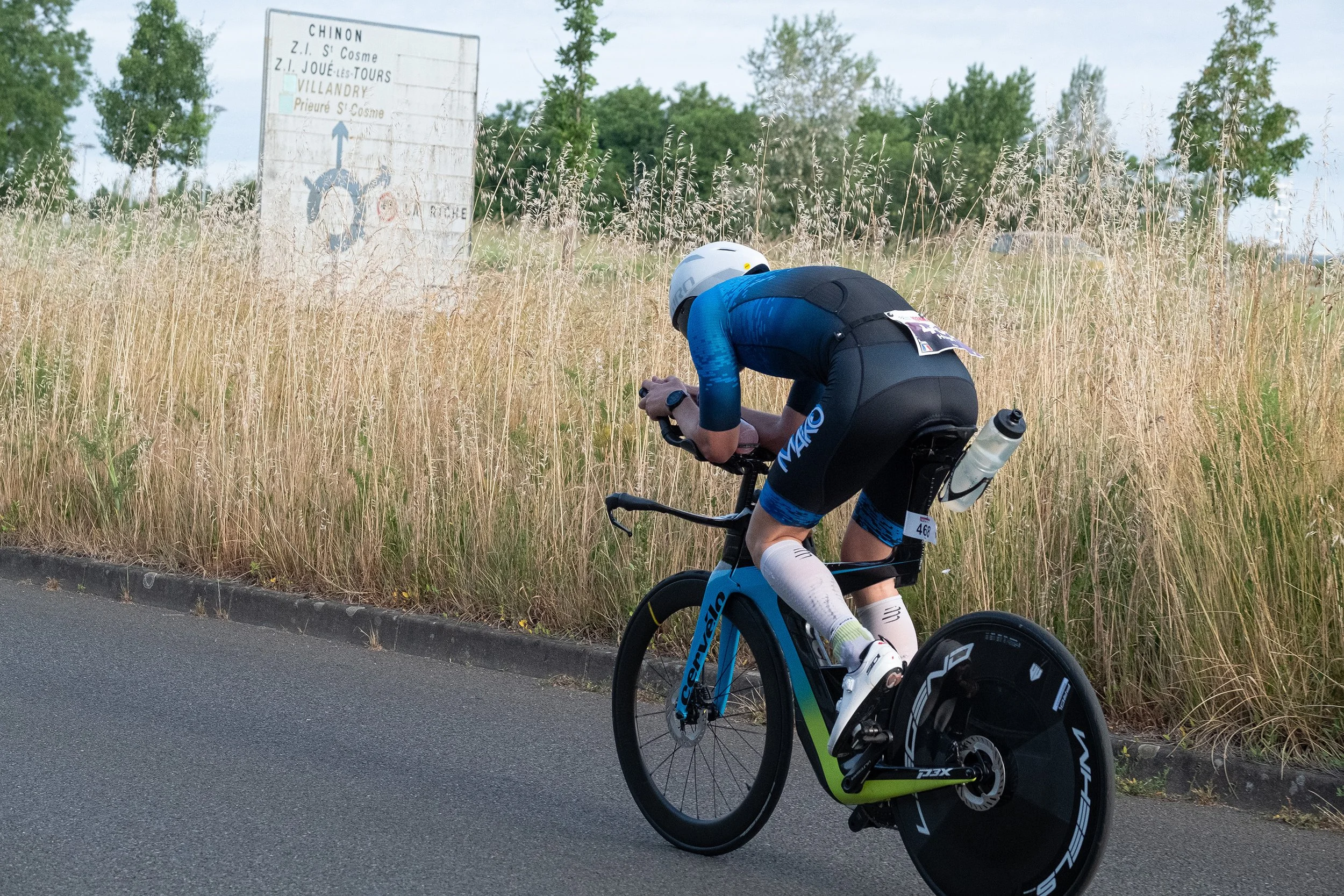 Un cycliste en tenue de course, portant un casque blanc, pédale lors d'une compétition sur une route asphaltée, avec de hautes herbes et un panneau indicateur de village en arrière-plan.