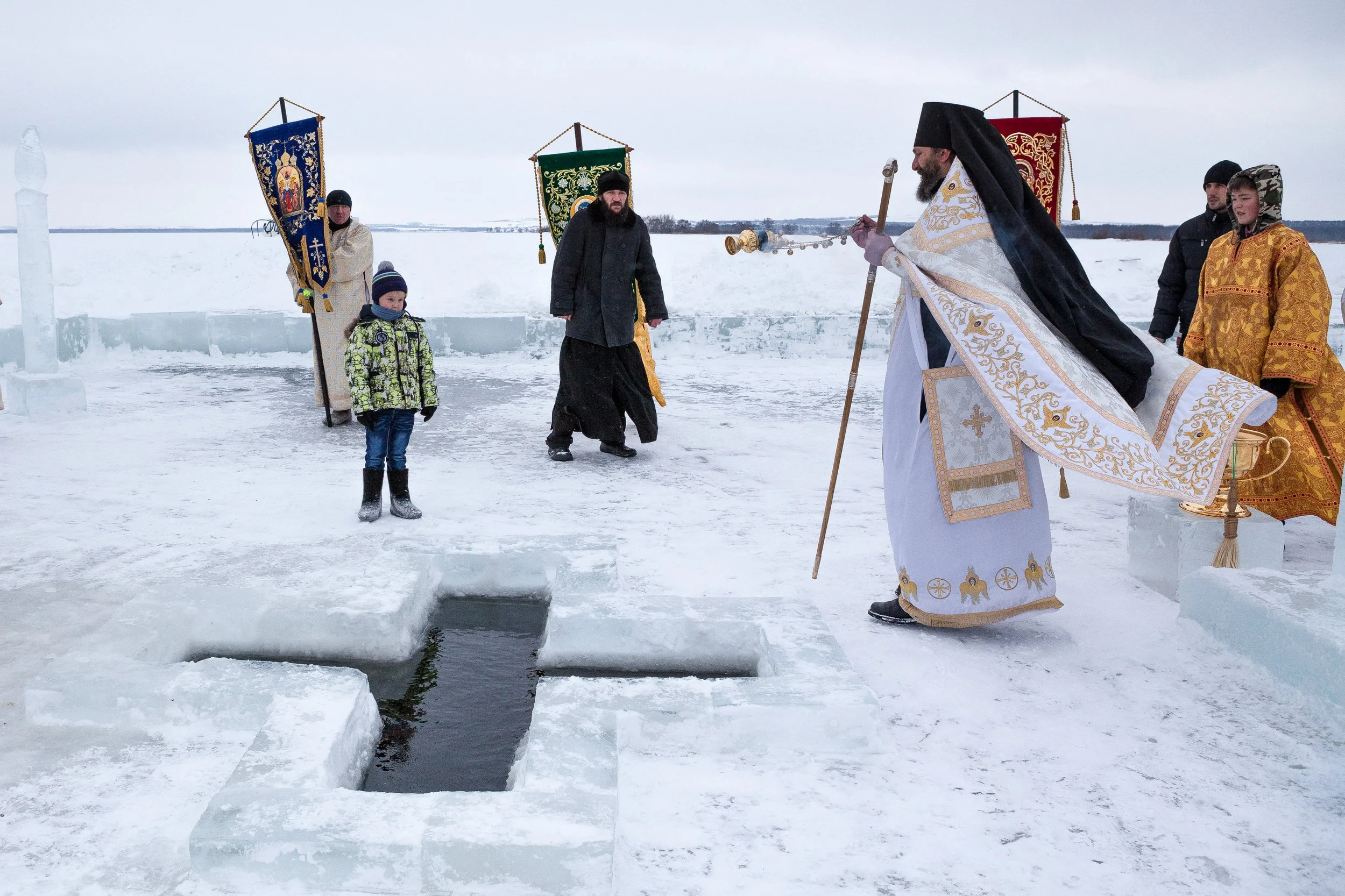 Religieux orthodoxes effectuant un rituel sur la glace, lors de l'épiphanie orthodoxe, avec une personne jeune observant, en milieu enneigé.