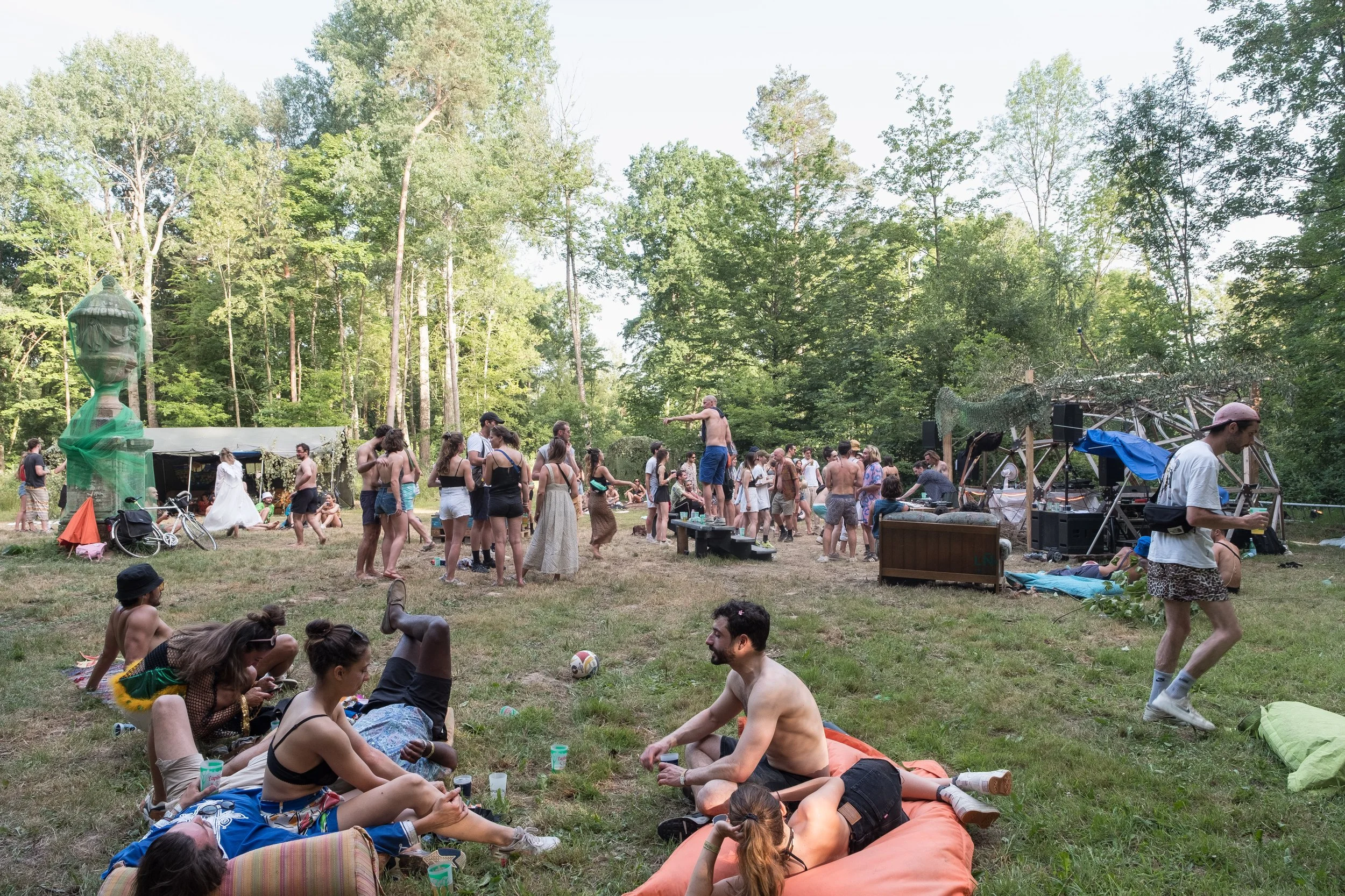 Fête en plein air dans une forêt avec des gens se relaxant, dansant, et partageant un moment convivial, avec une scène de musique et des structures en bois.
