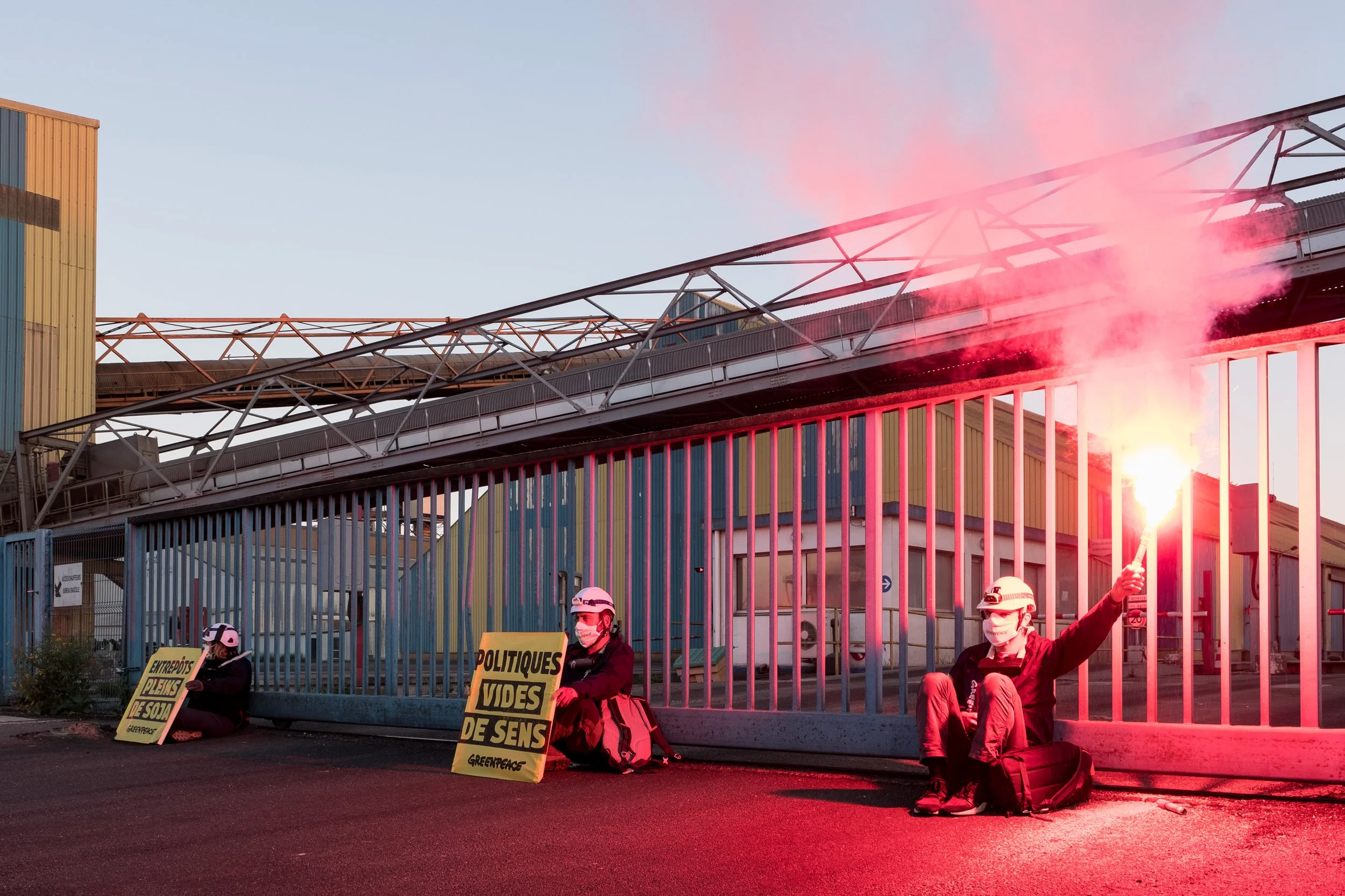 Trois activistes de Greenpeace assis devant un portail métallique, portant des casques et des masques, tenant des pancartes avec des messages écologiques, dont l'un affiche 'POLITIQUES VIDES DE SENS', et une personne tient un fumigène rouge.