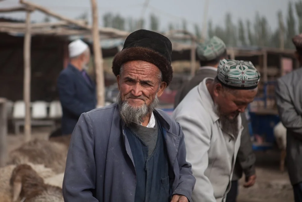 Groupe de personnes dans un marché, hommes portant des chapeaux traditionnels, un homme âgé avec une barbe grise en premier plan à Kashgar, en Chine.