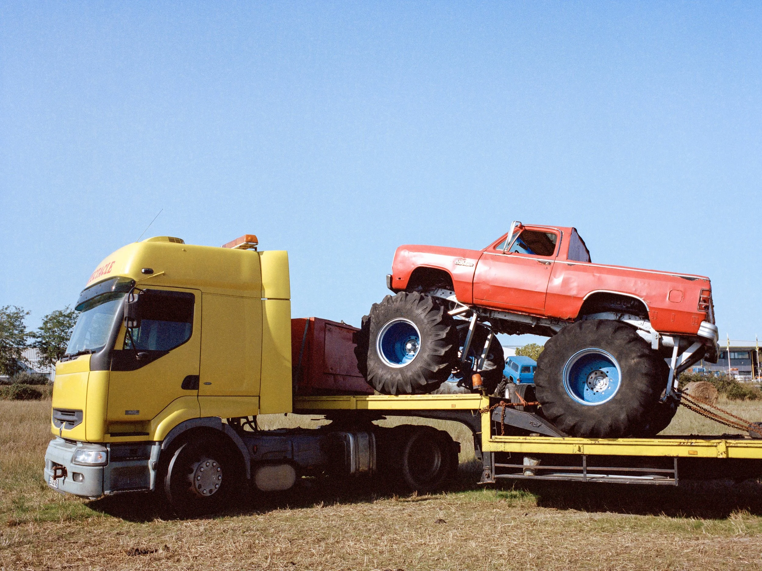 Camion jaune transportant unmonster truck rouge avec de grandes roues noires, ciel clair.