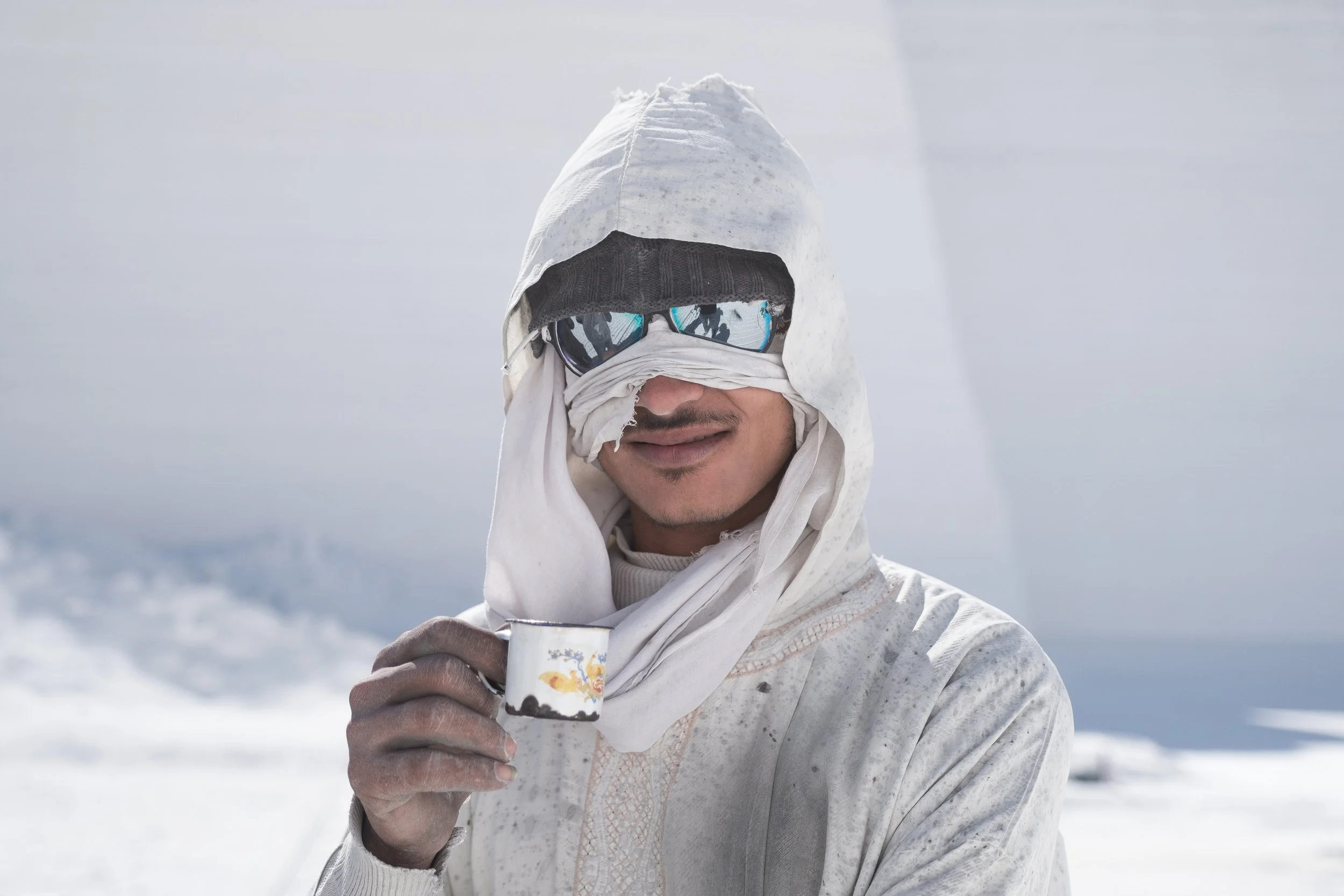 Homme portant des vêtements blancs et des lunettes de soleil dans un environnement tout blanc, tenant une tasse.