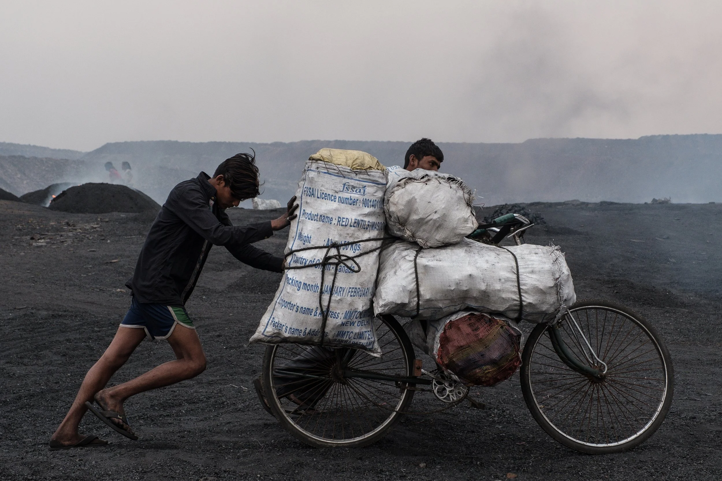 Deux jeunes hommes poussent un vélo chargé de sacs volumineux pleins de charbon dans un paysage de mouvements de fumée et de terrains volcaniques ou poussiéreux.