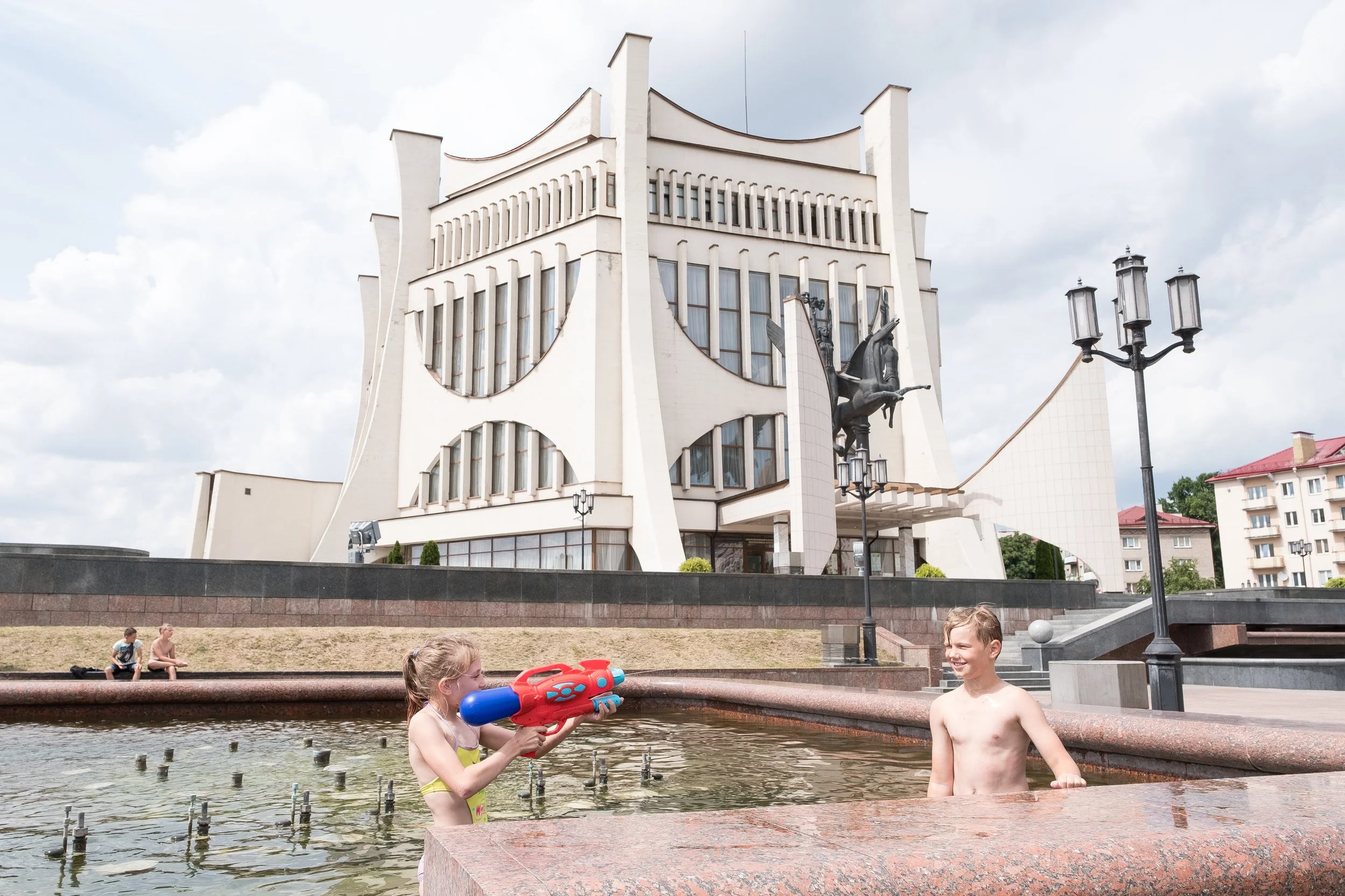 Deux enfants jouent avec un pistolet à eau dans une fontaine devant un bâtiment moderne blanc, avec une sculpture représentant un cheval ailé en bronze.