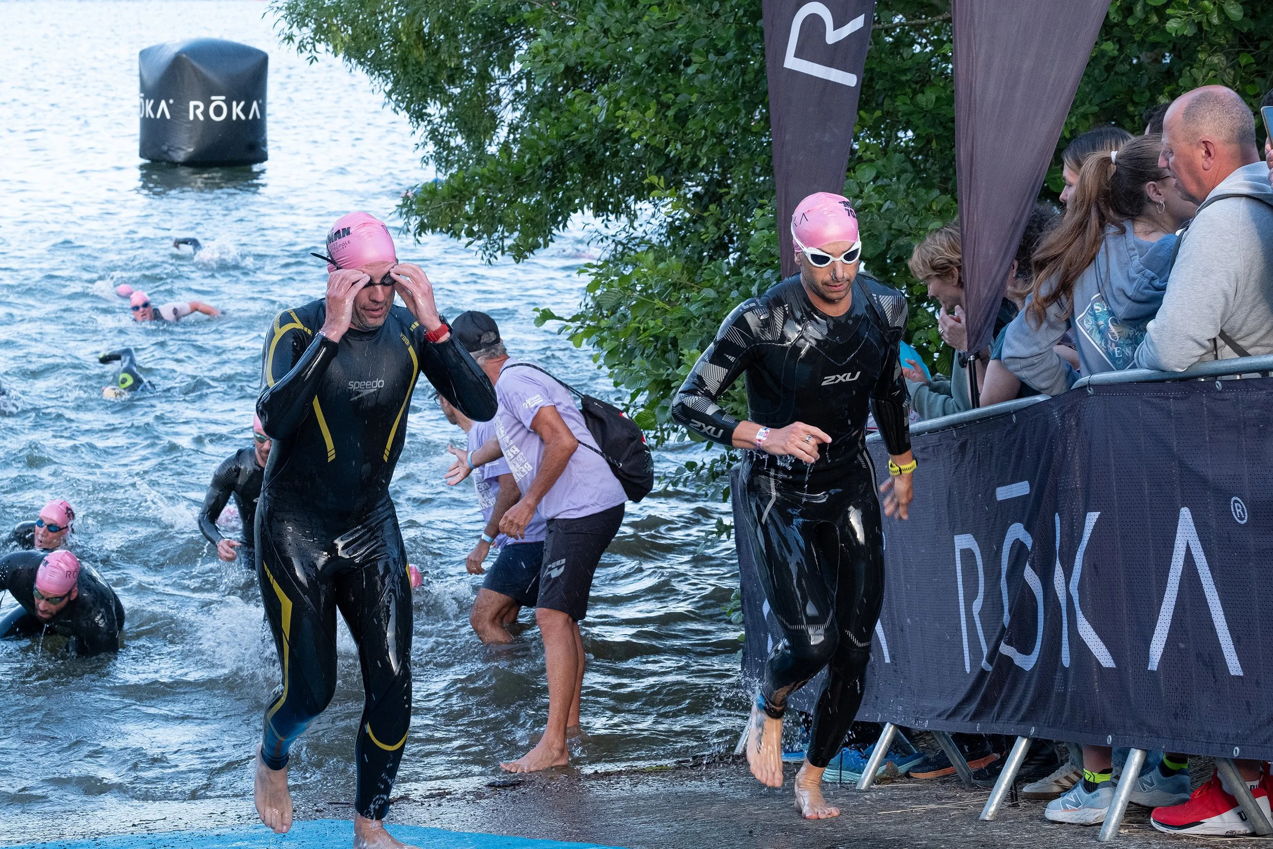 Participants sortis de l'eau lors d'une compétition de triathlon, portant des combinaisons de plongée, à côté d'une barrière avec un logo ROKA, au bord d'un plan d'eau entouré de végétation.