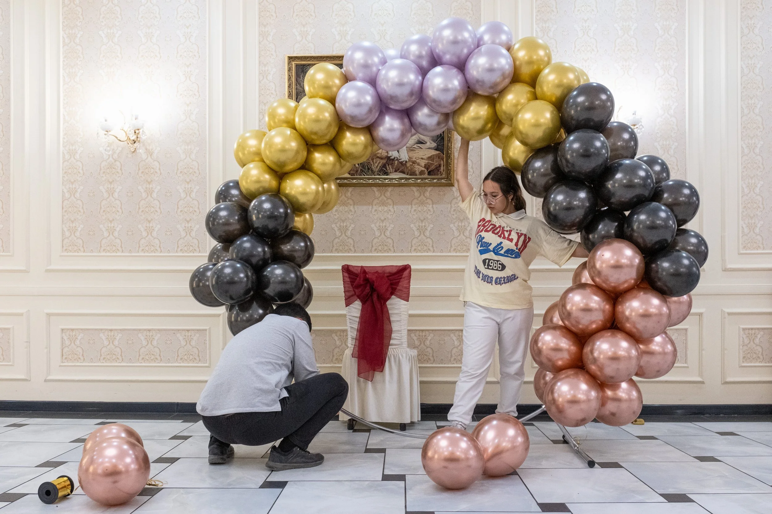 Deux personnes installent un décor de ballons pour une fête ou un événement dans une salle élégante, au Kazakhstan. L'un est accroupi, l'autre se tient debout, entourés de ballons roses, noirs, dorés et violets formant un arc.