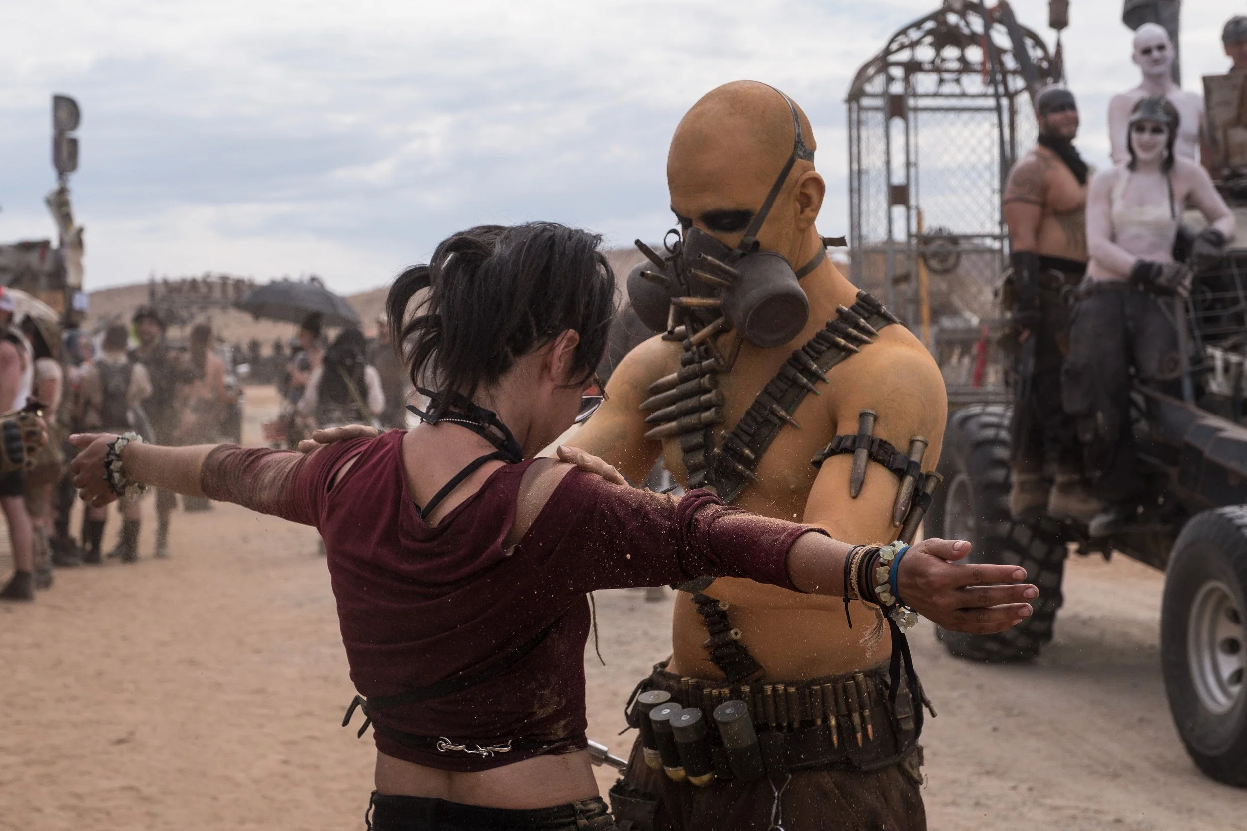 Deux personnes en costume post-apocalyptique dans un désert, l'une avec un masque à gaz, l'autre avec un maquillage et des accessoires de style futuriste, dans un paysage désertique avec d'autres personnes en arrière-plan.