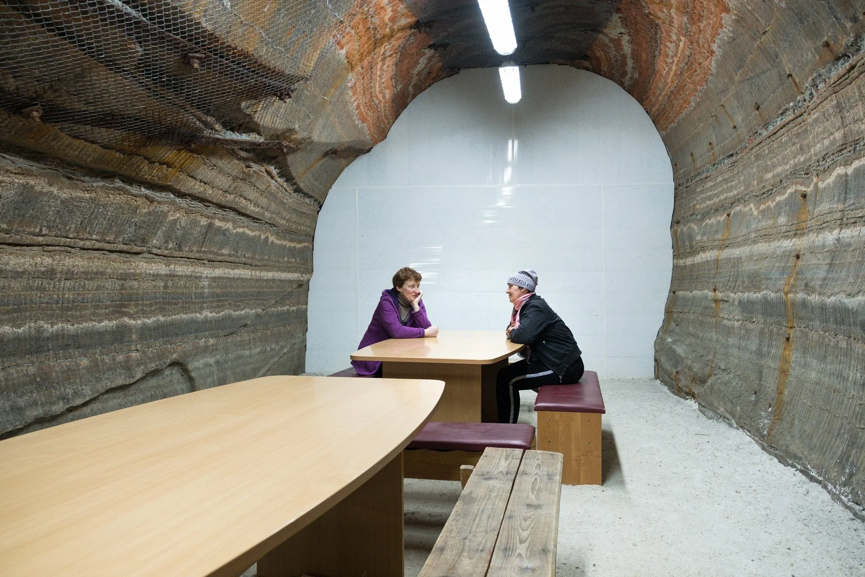 Deux femmes discutent assises à une table dans une pièce souterraine avec des murs et un plafond en roche. Un mur blanc est à l'arrière-plan. La pièce est éclairée par deux tubes fluorescents.