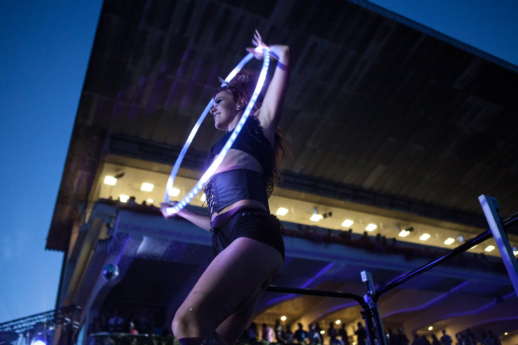 Une jeune femme danse avec un hula hoop lumineux lors d'une performance en soirée devant un public. Elle porte un haut noir pailleté et un short noir, avec un ciel nocturne.