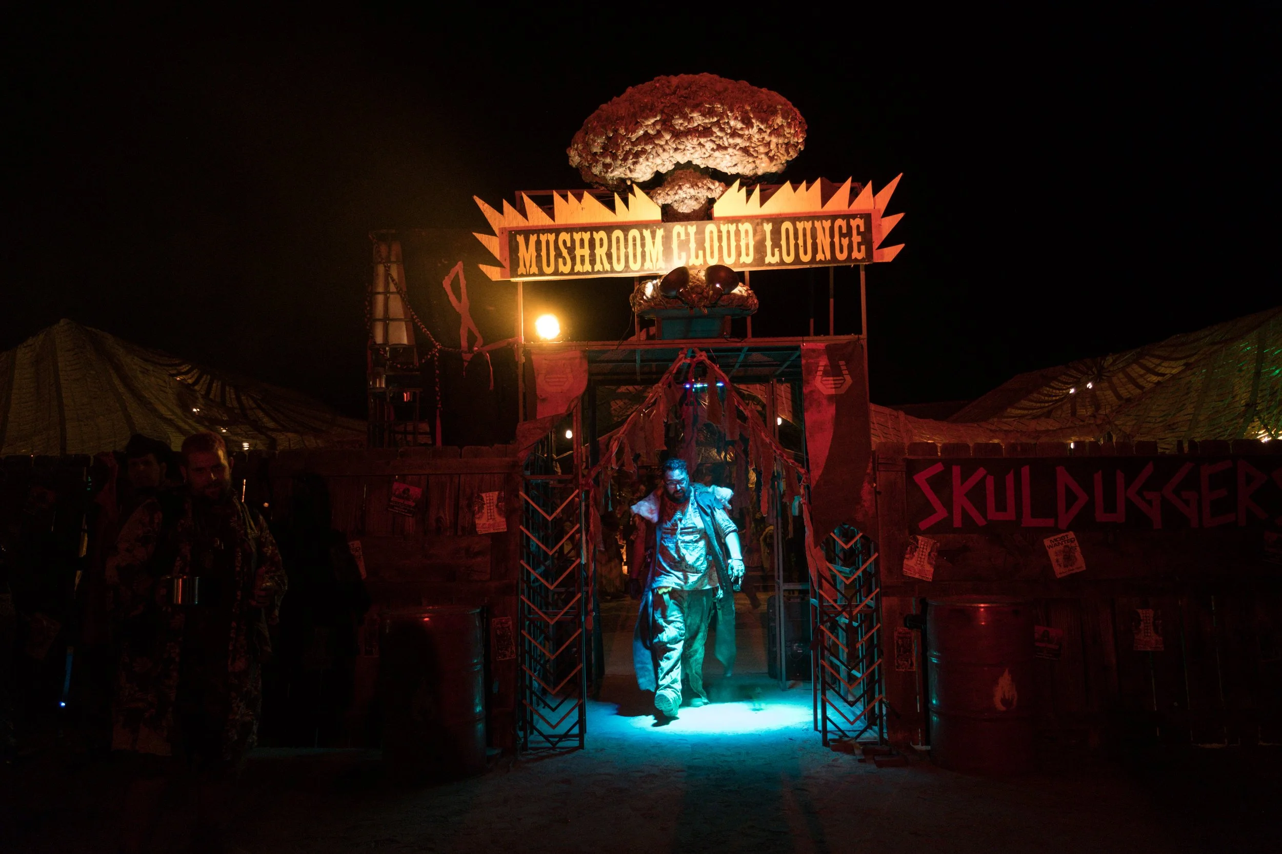 Entrée du Mushroom Cloud Lounge au festival Wasteland, avec un homme avançant dans une lumière bleue. Le décor est sombre avec un éclairage rouge et une décoration inspirée d'une explosion nucléaire.