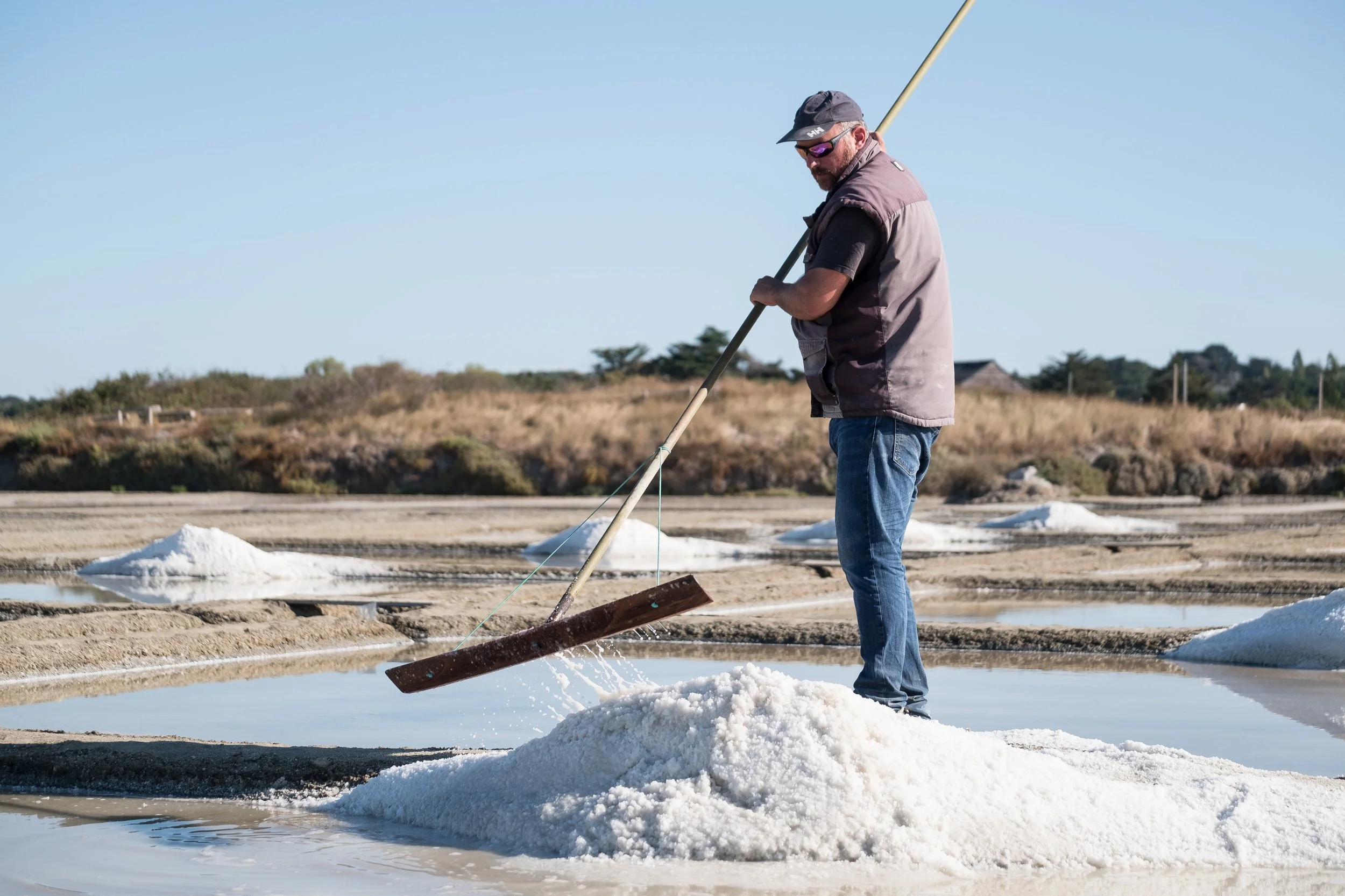 Homme récoltant du sel dans une saline à ciel ouvert, utilisant un râteau pour ramasser le sel.