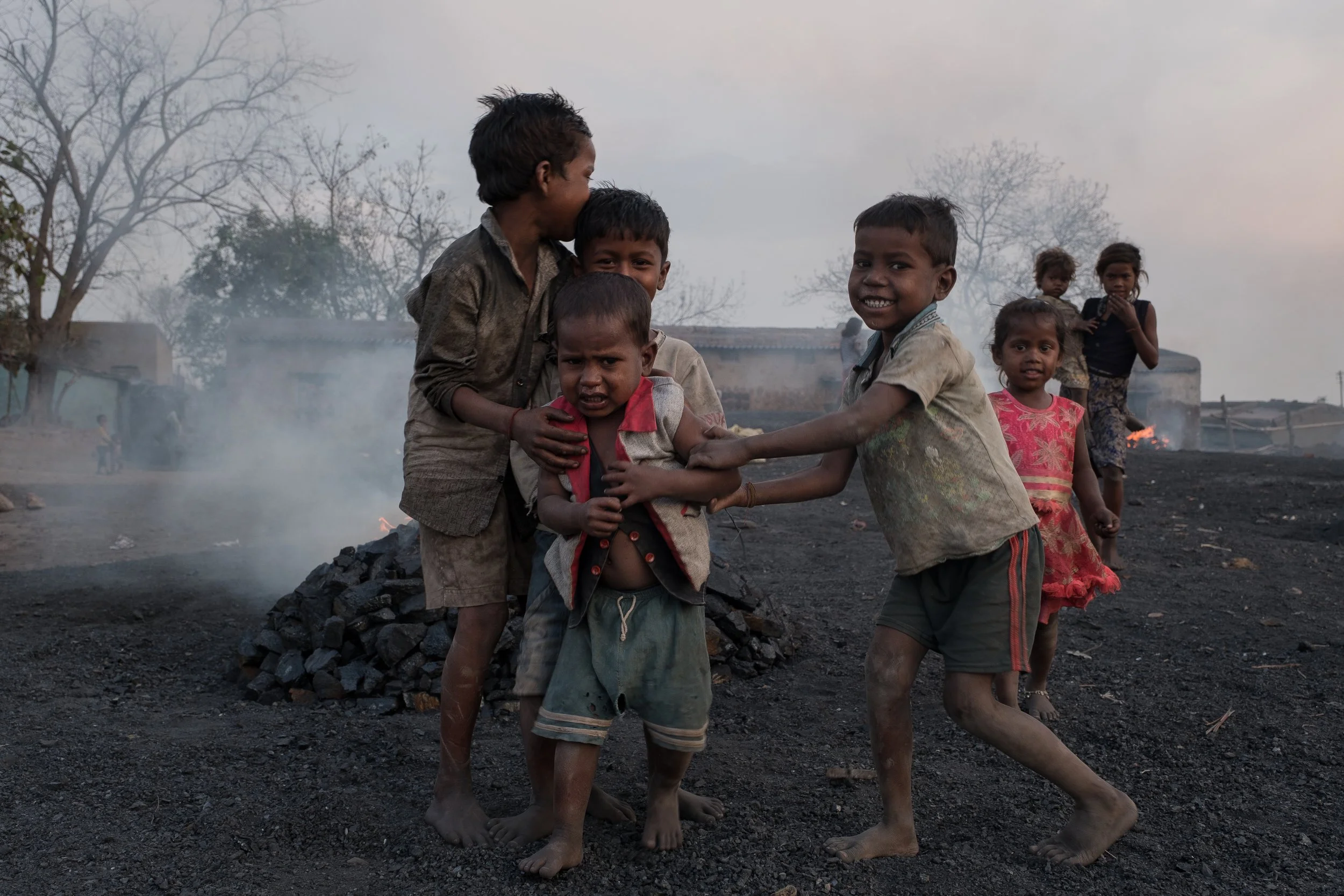 Des enfants jouent sur un terrain noir de charbon avec de la fumée et des bâtiments en arrière-plan