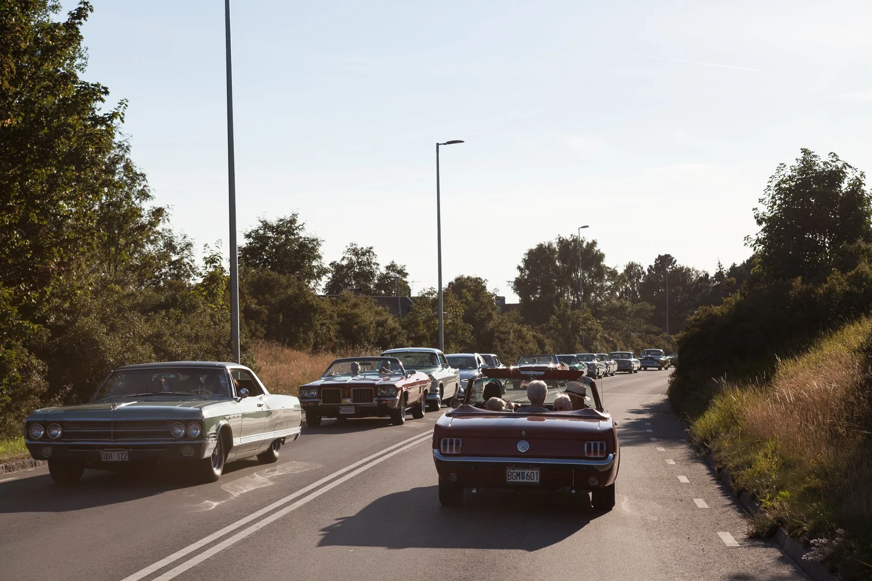 Une procession de voitures classiques, dont un cabriolet rouge avec des passagers, circule sur une route bordée d'arbres et de lampadaires sous un ciel ensoleillé en Suède.
