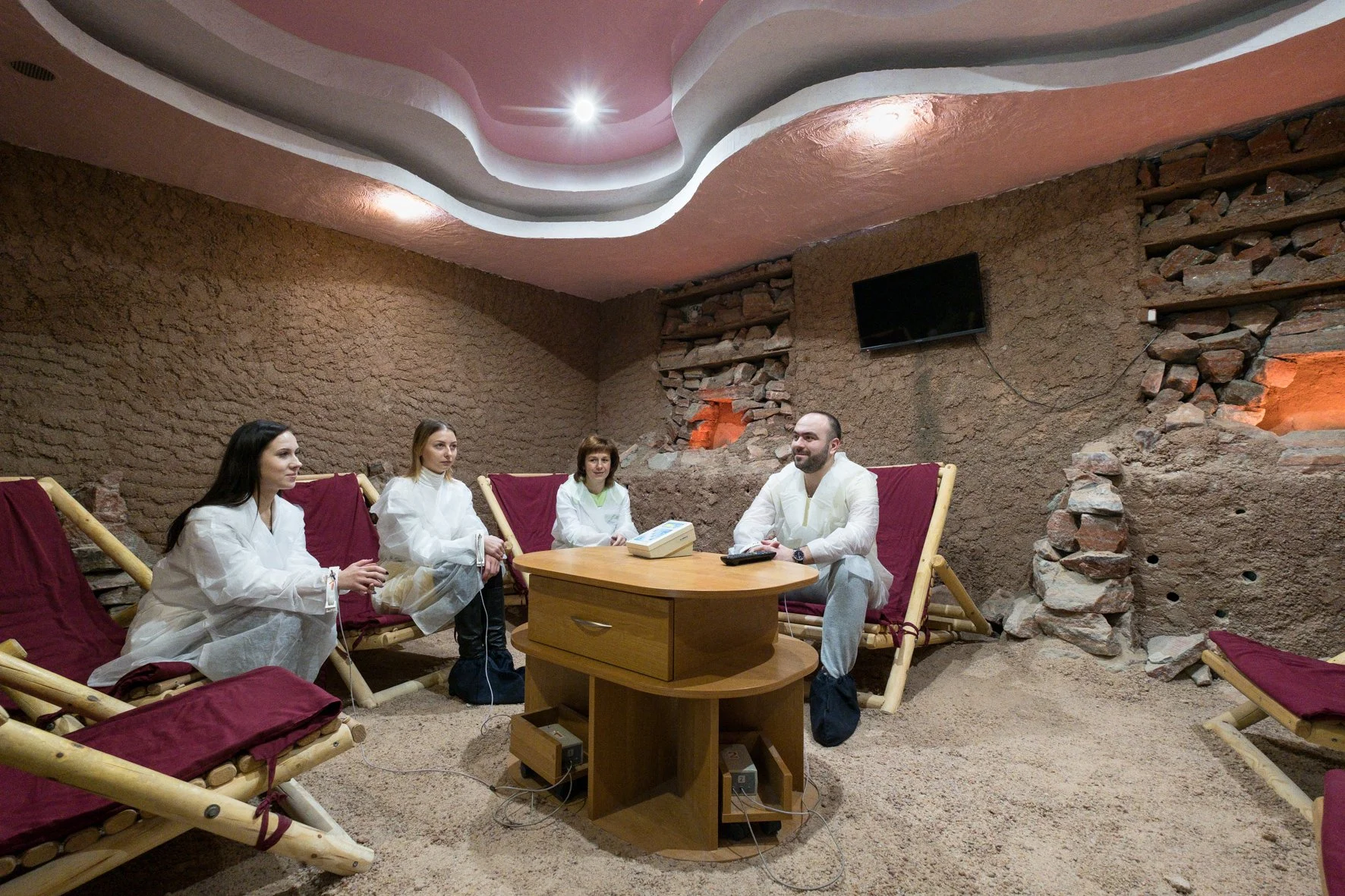 Un groupe de personnes assises dans une salle de sanatorium, avec des murs en terre cuite et un plafond peint en rose et gris, des bancs en bois avec des coussins rouges, un petit meuble avec des appareils électroniques.