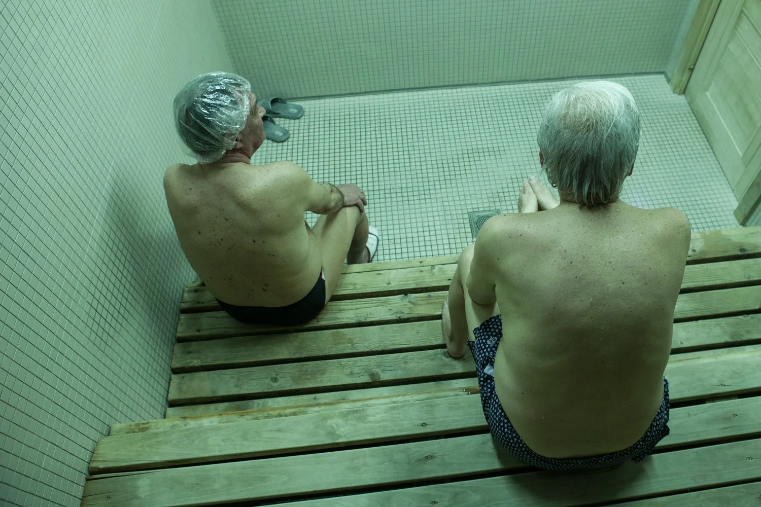Deux personnes âgées assises dans un sauna, vue du dessus, avec des murs en carreaux et un sol en mosaïque, portant des coiffes de douche jetables.