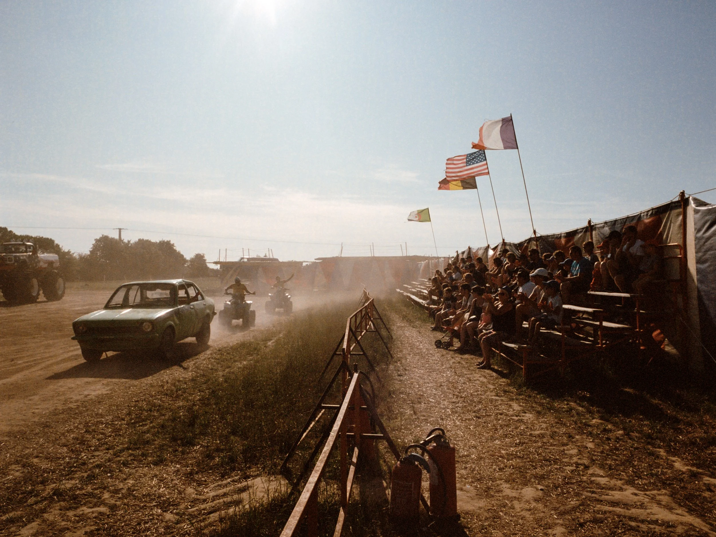 Spectateurs assis sur des gradins en plein air regardant un défilé de voitures sur un terrain poussiéreux, avec des drapeaux flottant dans le vent et un ciel ensoleillé.