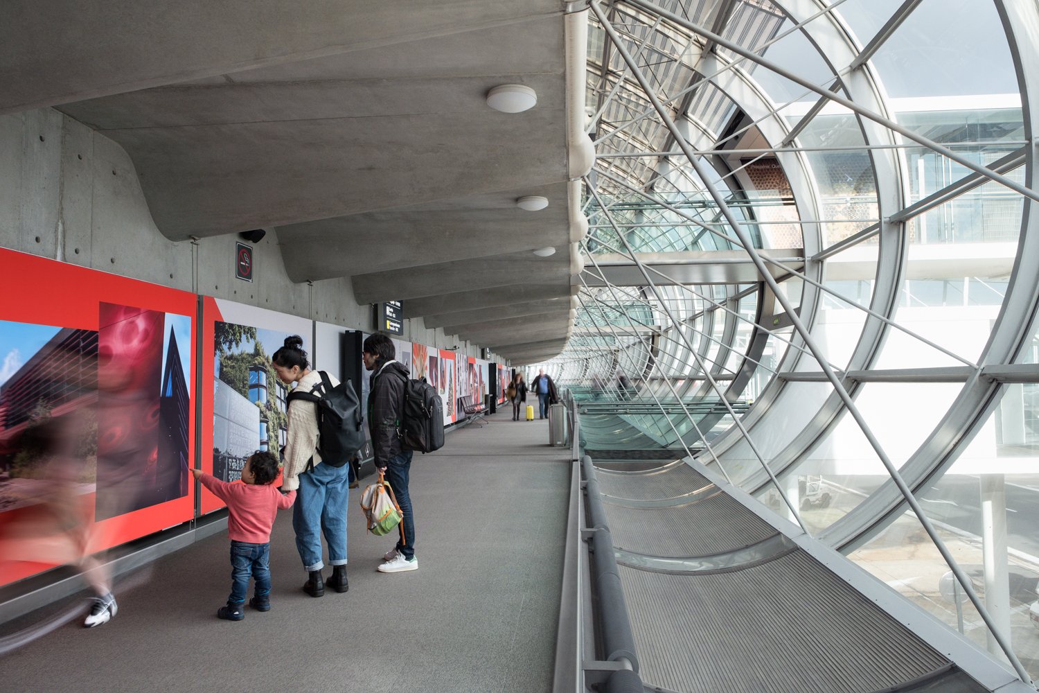 Des personnes, dont une famille avec un petit enfant, regardent une exposition à l'intérieur de l'aéroport Charles de Gaulle avec de grandes fenêtres incurvées en verre.