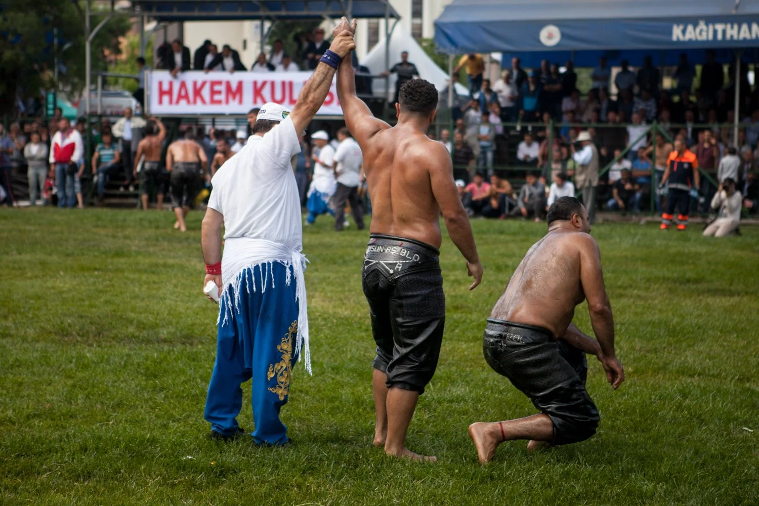 Trois hommes, deux athlètes et un arbitre, à la fin d'un combat de lutte en plein air, avec un groupe de personnes derrière eux. Un des hommes célèbre sa victoire en saluant la foule main levée, lors d'une compétition.