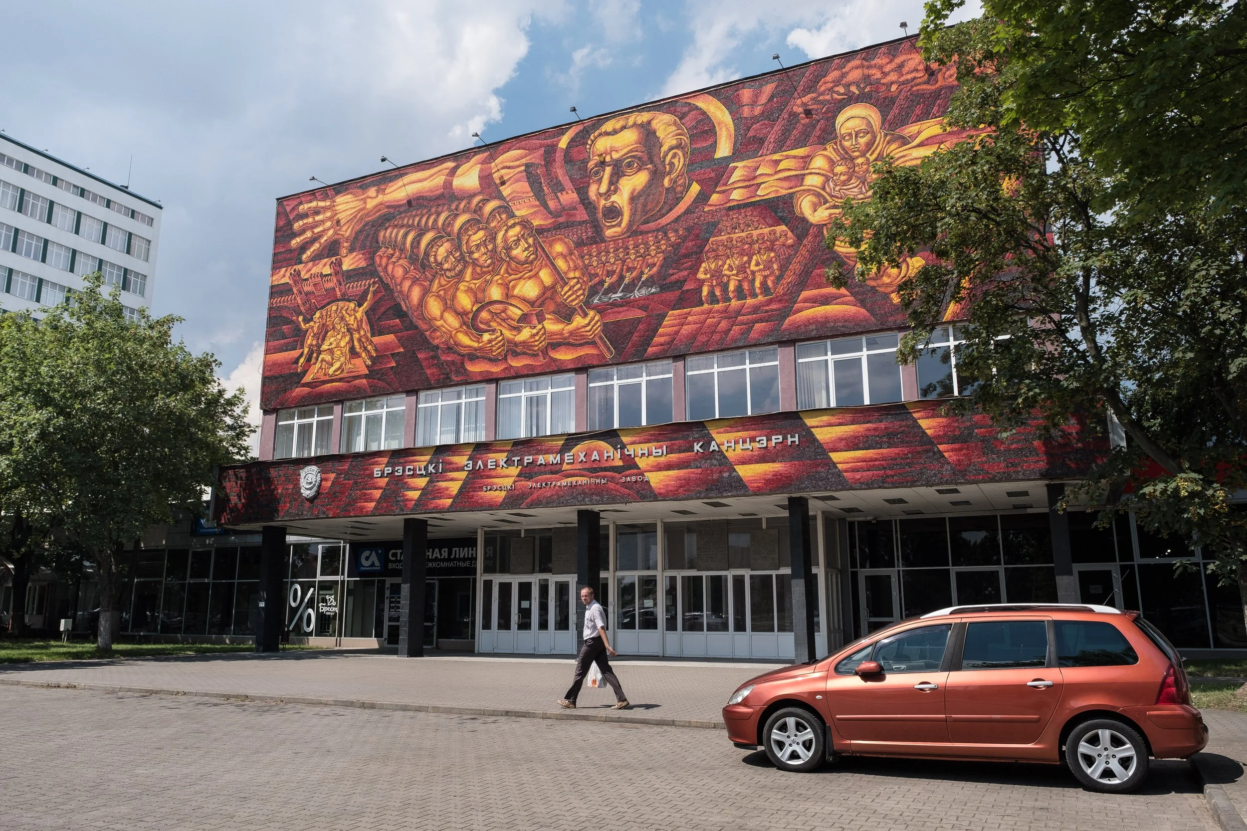 Bâtiment avec une grande fresque murale soviétique représentant des figures symboliques, et des inscriptions en cyrillique sur la façade, avec un homme marchant et une voiture orange stationnée devant en Biélorussie.