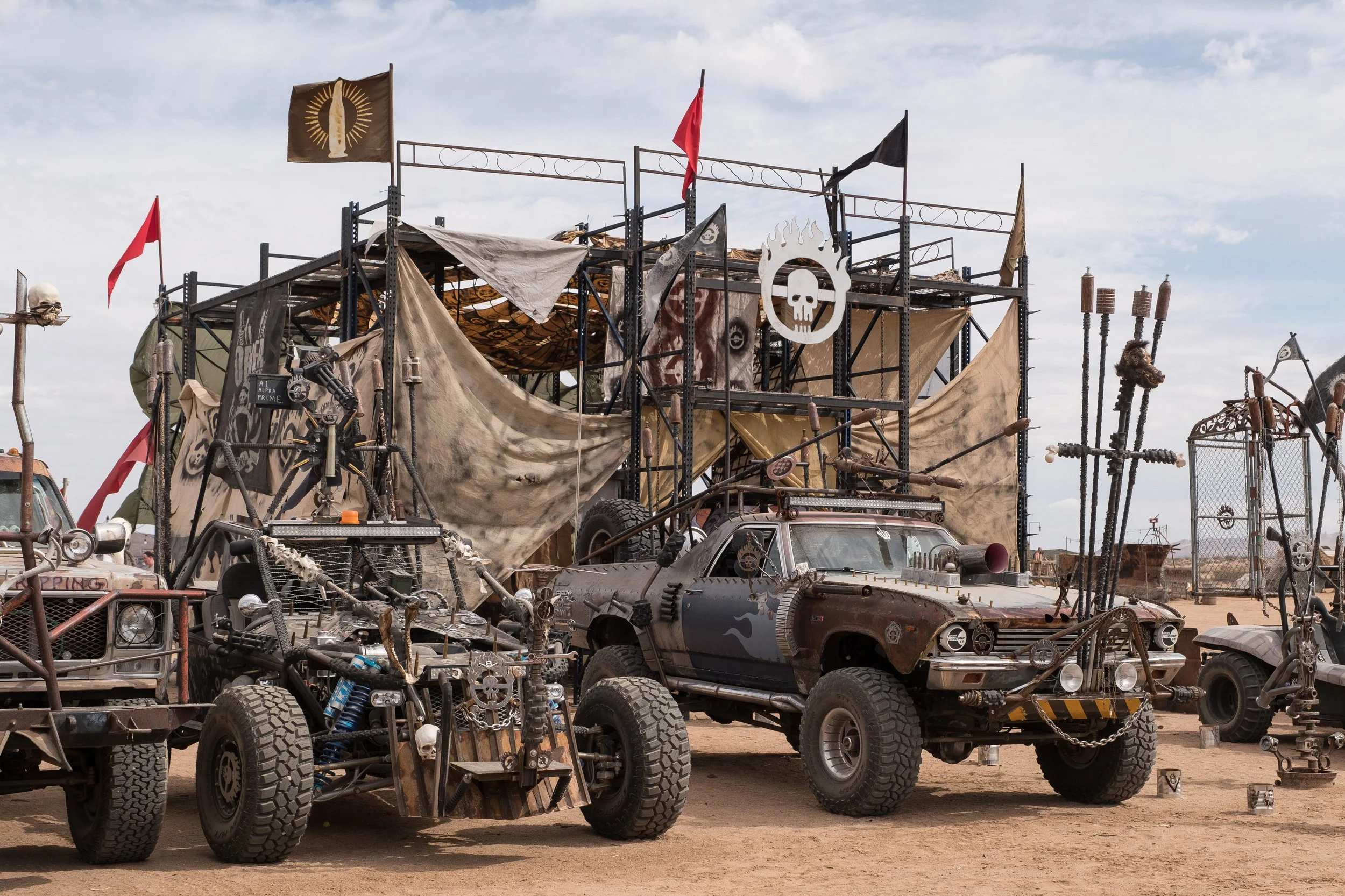 Une scène de voitures et structures en métal rouillés, décorées avec des drapeaux, des crânes, des chaînes et des accessoires dans un désert, évoquant un univers dystopique ou post-apocalyptique à Wasteland.