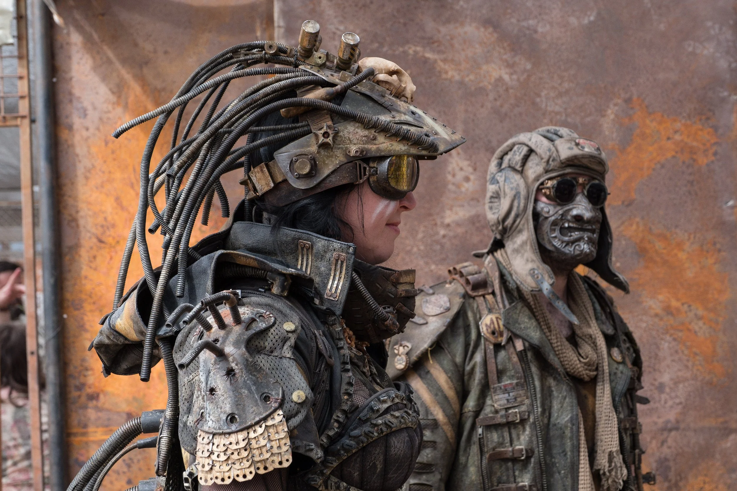 Deux personnes en costumes futuristes et usés avec accessoires technologiques, l'une portant un casque avec câbles et une autre portant un masque, face à un mur rouillé à Wasteland.
