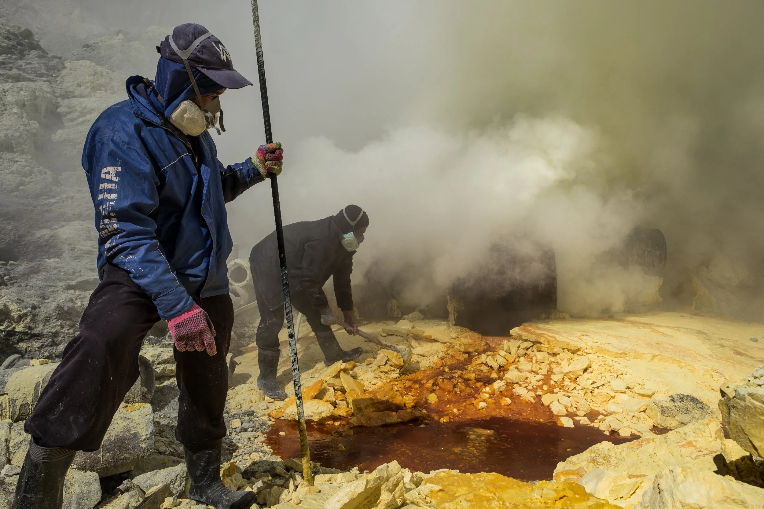 Deux mineurs portant des équipements de protection travaillant dans un environnement de vapeur et de roches volcaniques, manipulent du sulfure rougeâtre extrêmement chaud au sol.