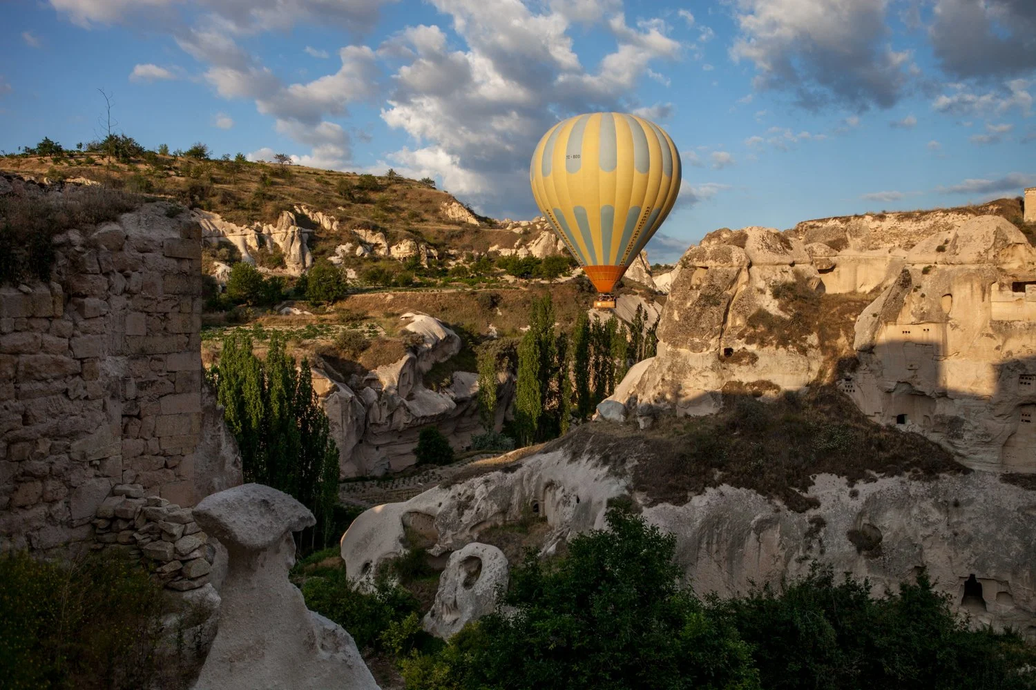 Montgolfière jaune volant au-dessus d'un paysage rocheux avec des formations géologiques uniques et des arbres dans une vallée au-dessous, au lever du soleil.