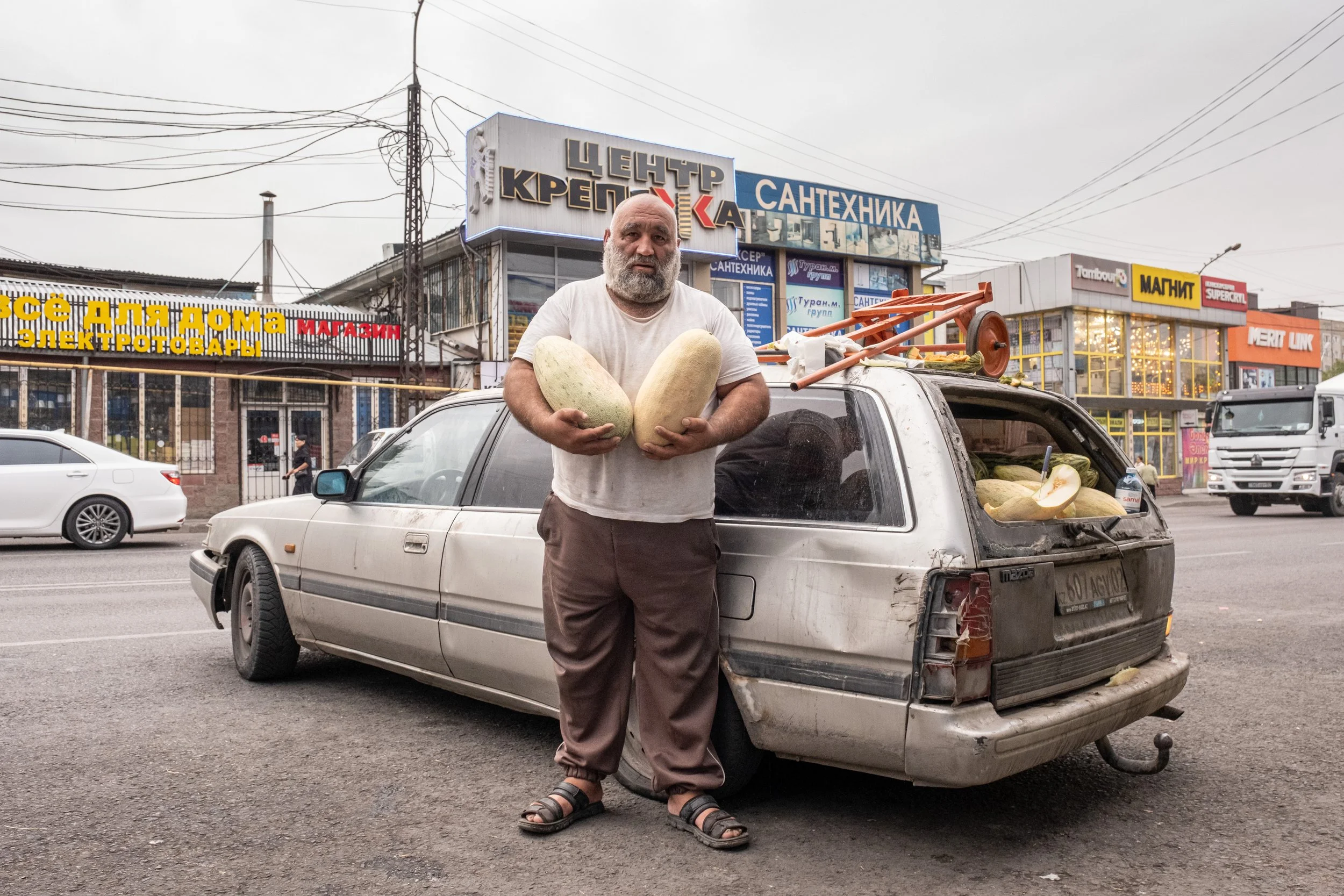 Un homme debout devant une vieille voiture, tenant deux grands melons. La voiture est chargée de melons et stationne dans une rue urbaine avec des magasins en arrière-plan au Kazakhstan.