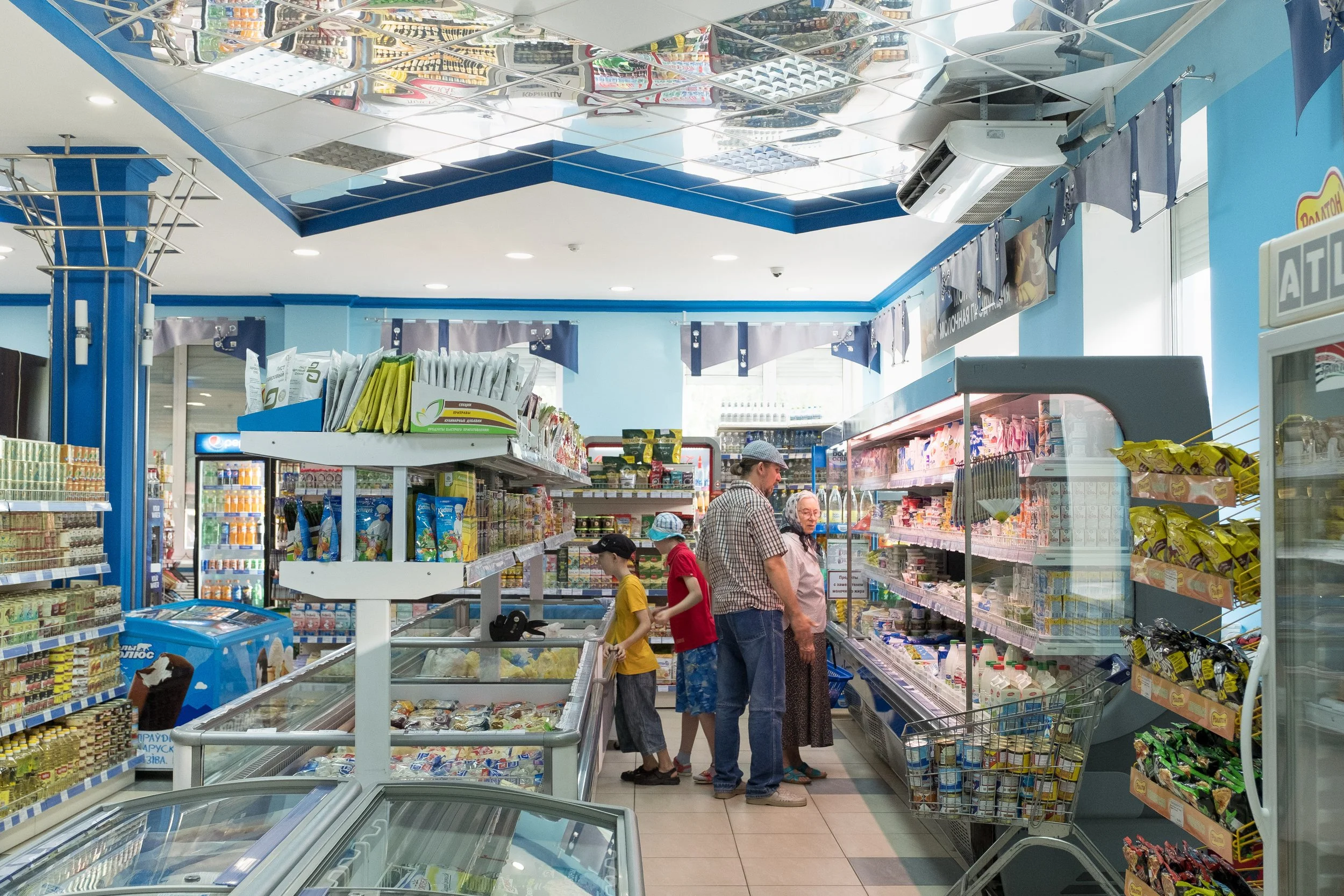 Intérieur d'une supérette avec des rayons de produits alimentaires. Quatre personnes, dont deux enfants, achètent des produits. La décoration est bleu clair avec des rayonnages bien organisés en Biélorussie.