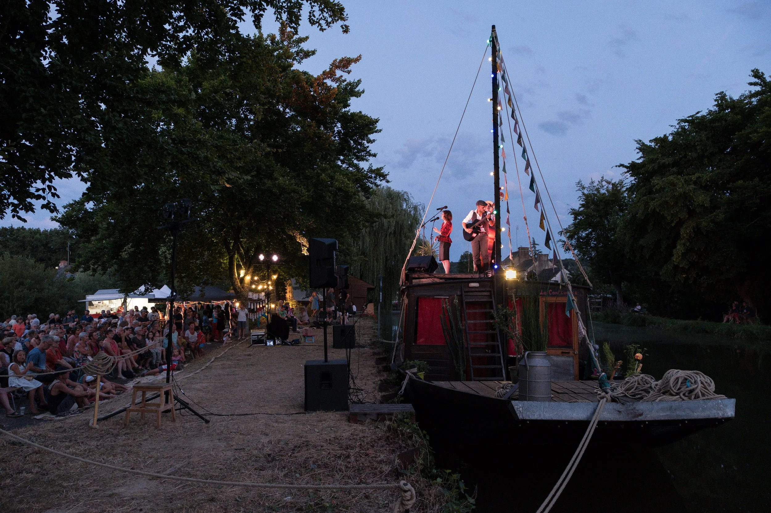 Spectacle en plein air sur un bateau en guise de scène, avec deux musiciens jouant, un public en soirée près d'un cours d'eau, éclairages décoratifs et arbres en arrière-plan.