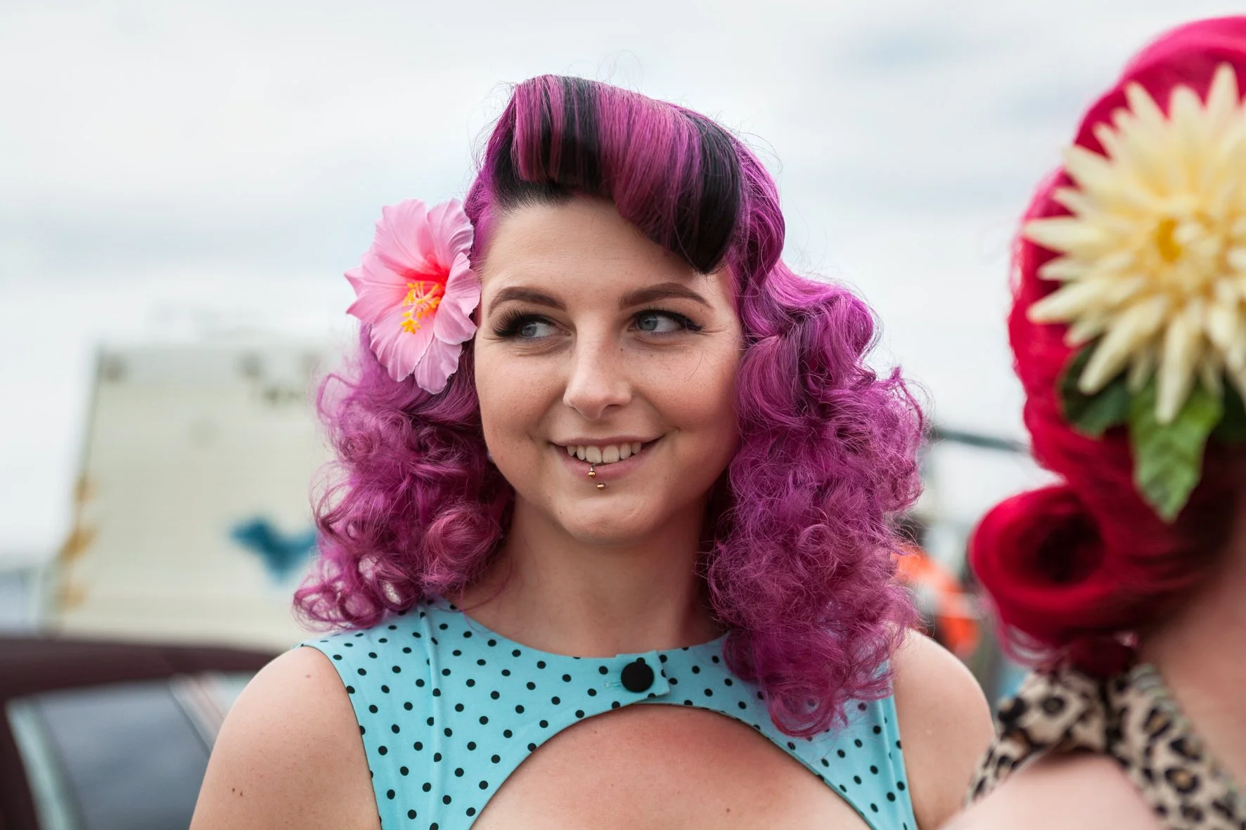 Femme pin-up avec cheveux bouclés et teints en violet, portant une fleur dans les cheveux, souriant.