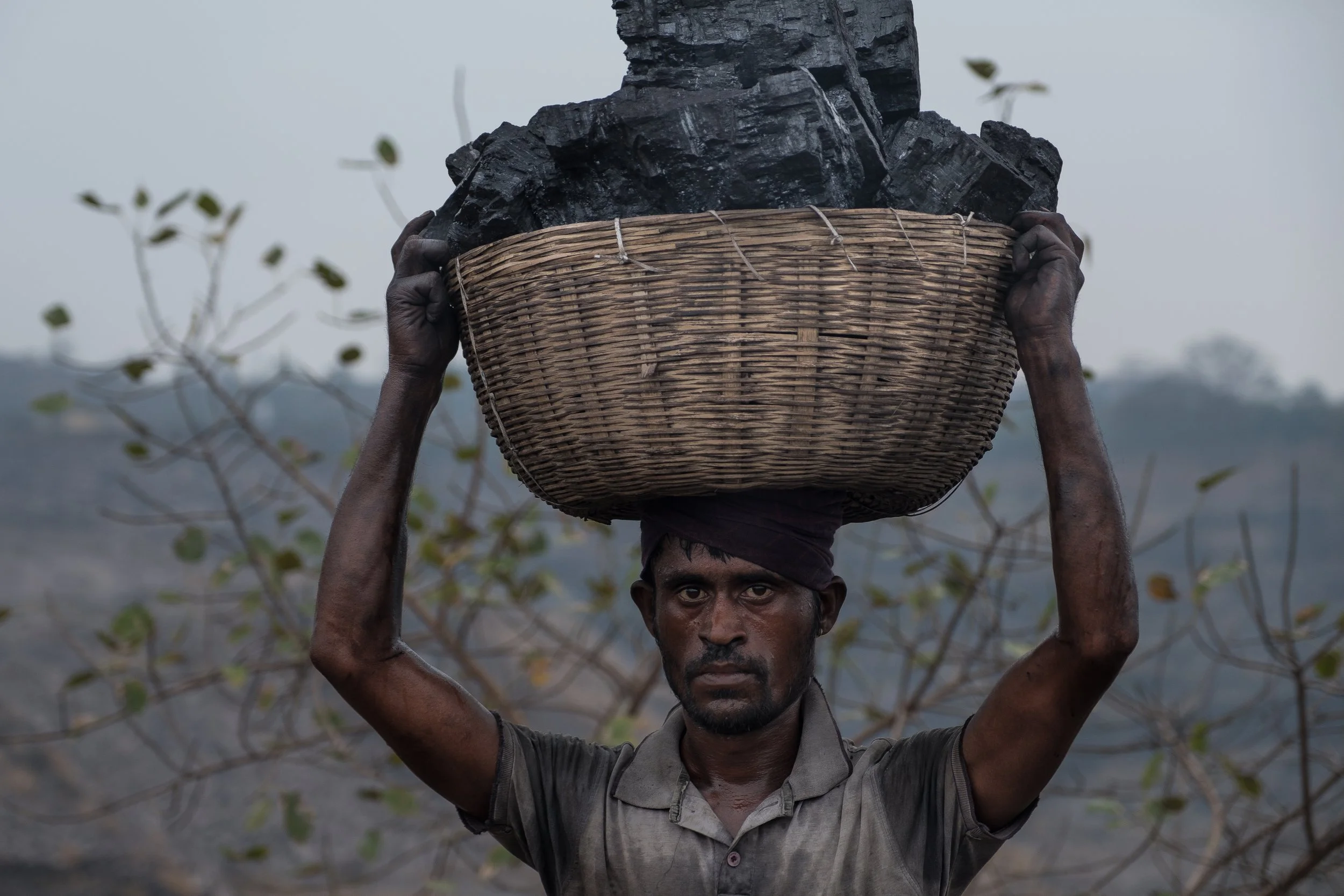 Un homme avec la peau sombre, portant un bandana noir, porte un grand panier rempli de charbon de bois sur sa tête, dans un paysage rural.