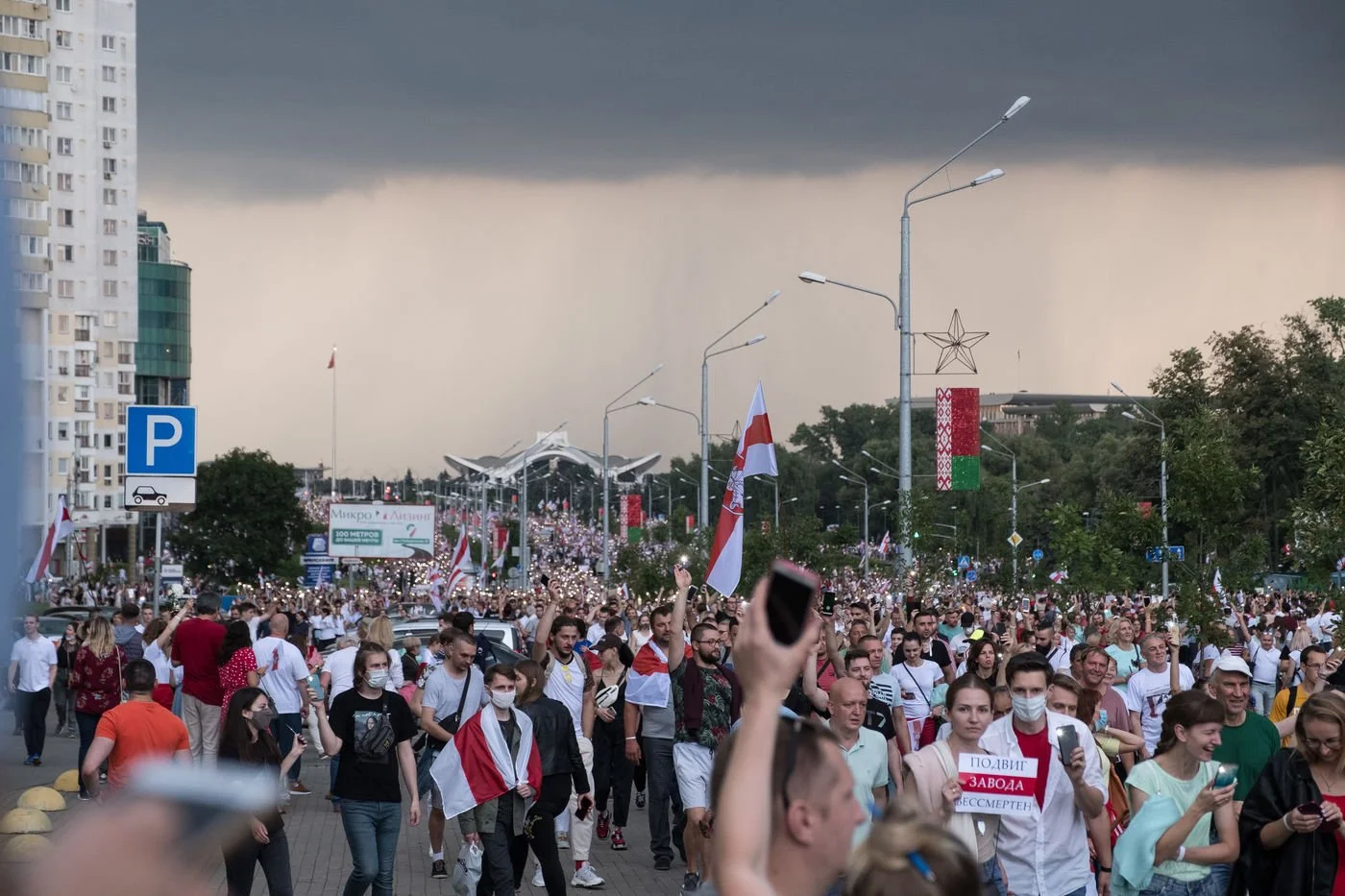 Une grande foule de manifestants portant des drapeaux blanc et rouge, certains portant des masques, manifeste dans une rue de Minsk en Biélorussie sous un ciel orageux, avec des bâtiments et des décorations en arrière-plan.