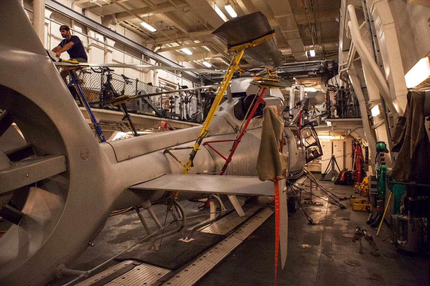 Intérieur d'une salle de la frégate militaire Surcouf, avec un hélicoptère en maintenance, des outils, des équipements et une personne travaillant sur le bateau.