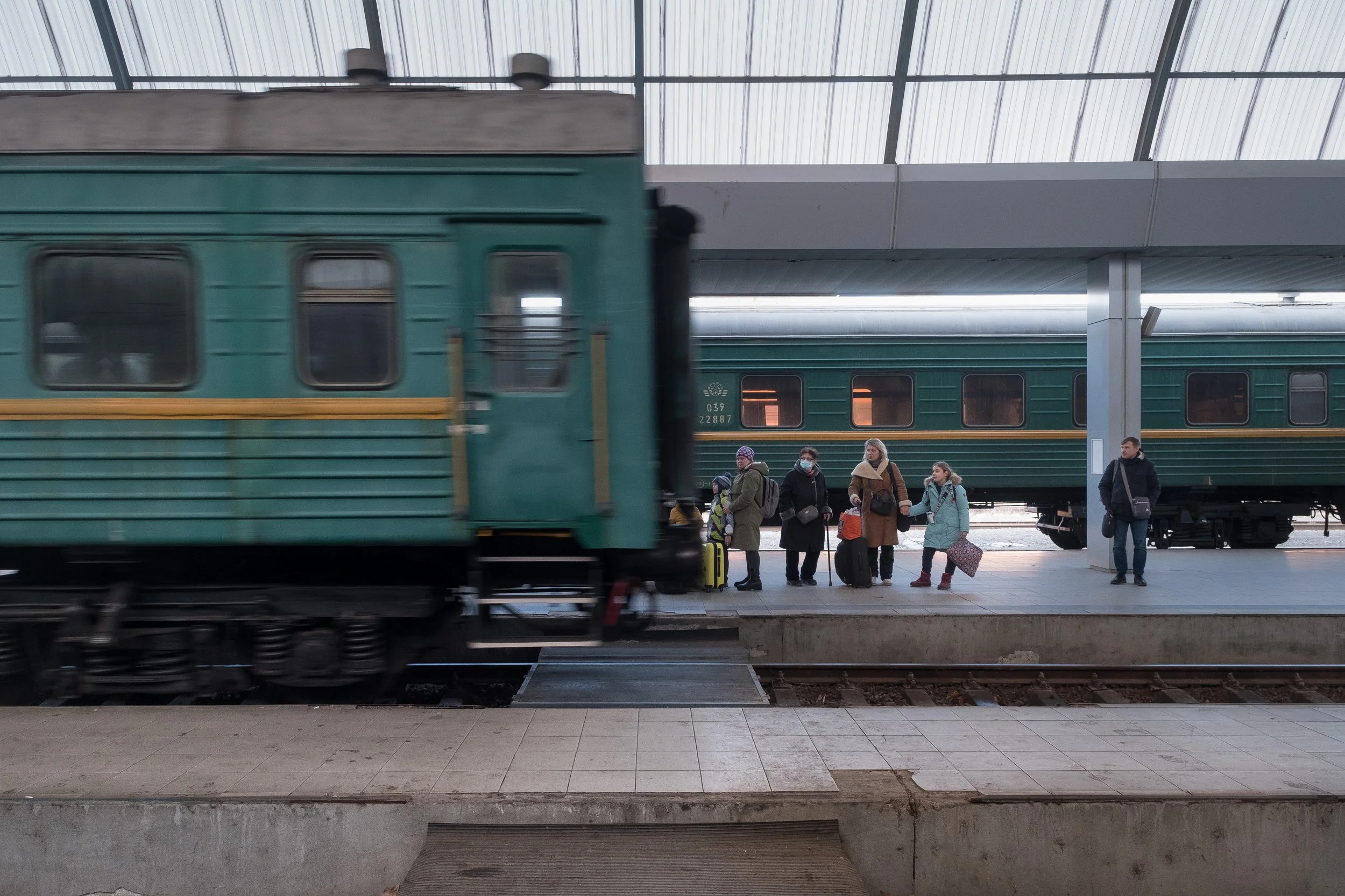 Des réfugiés avec des valises attendant à une gare ferroviaire, un train vert en mouvement passe devant eux.