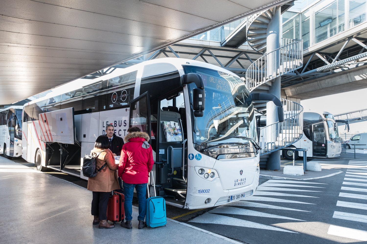 Groupe de trois personnes avec valises devant un bus blanc à l'aéroport, près d'une passerelle, préparation à l'embarquement.