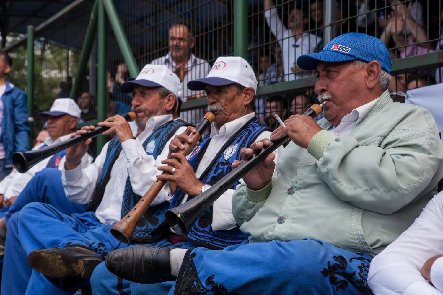Groupe de musiciens âgés jouant de la clarinette, portant des casquettes et des vestons bleus, lors d'une compétition de lutte, avec des spectateurs en arrière-plan.