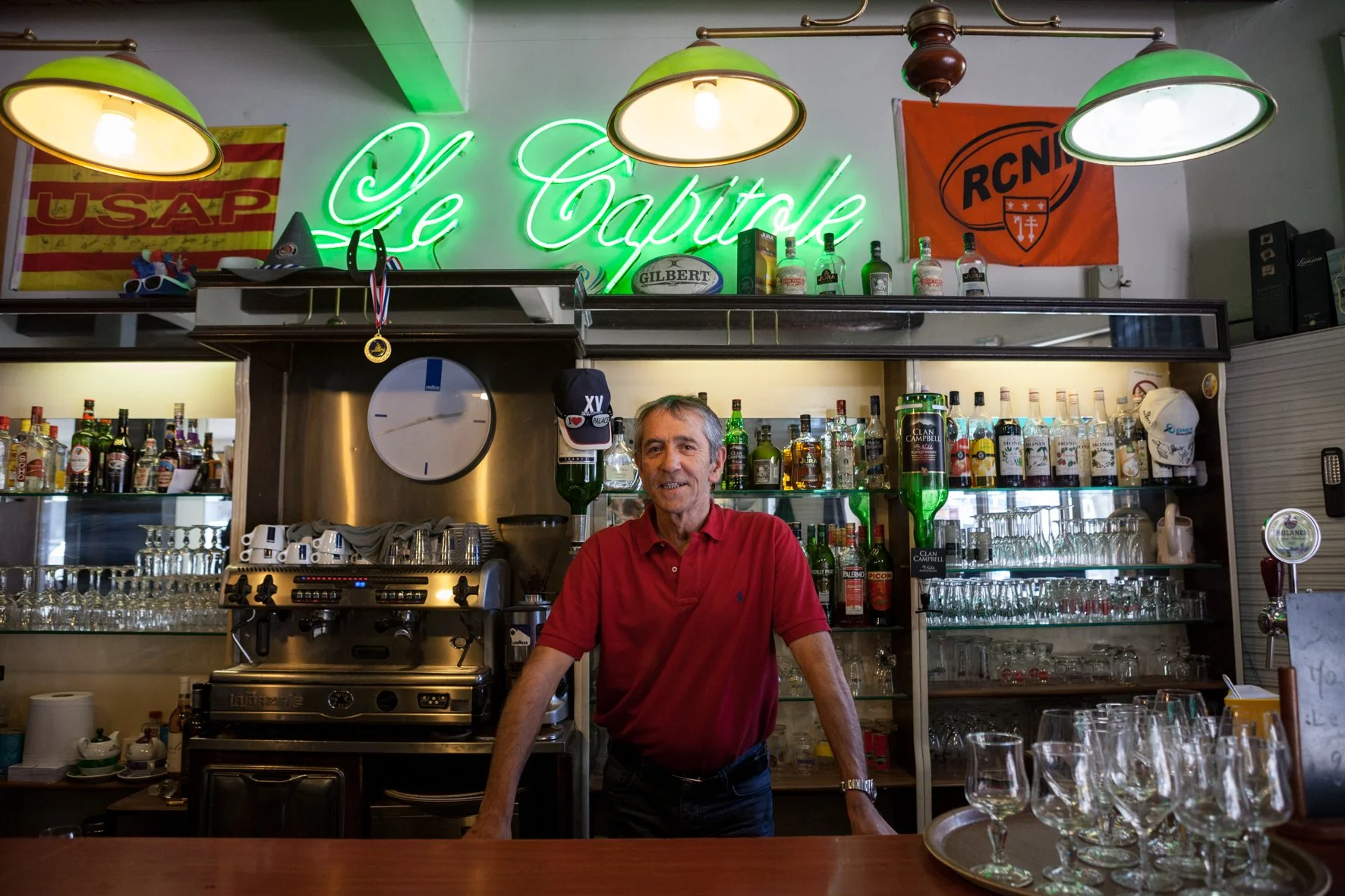Un homme souriant derrière un bar, entouré de bouteilles d'alcool, de verres et de matériel de bar, dans un établissement appelé "Le Capitole" avec un néon vert et des drapeaux.